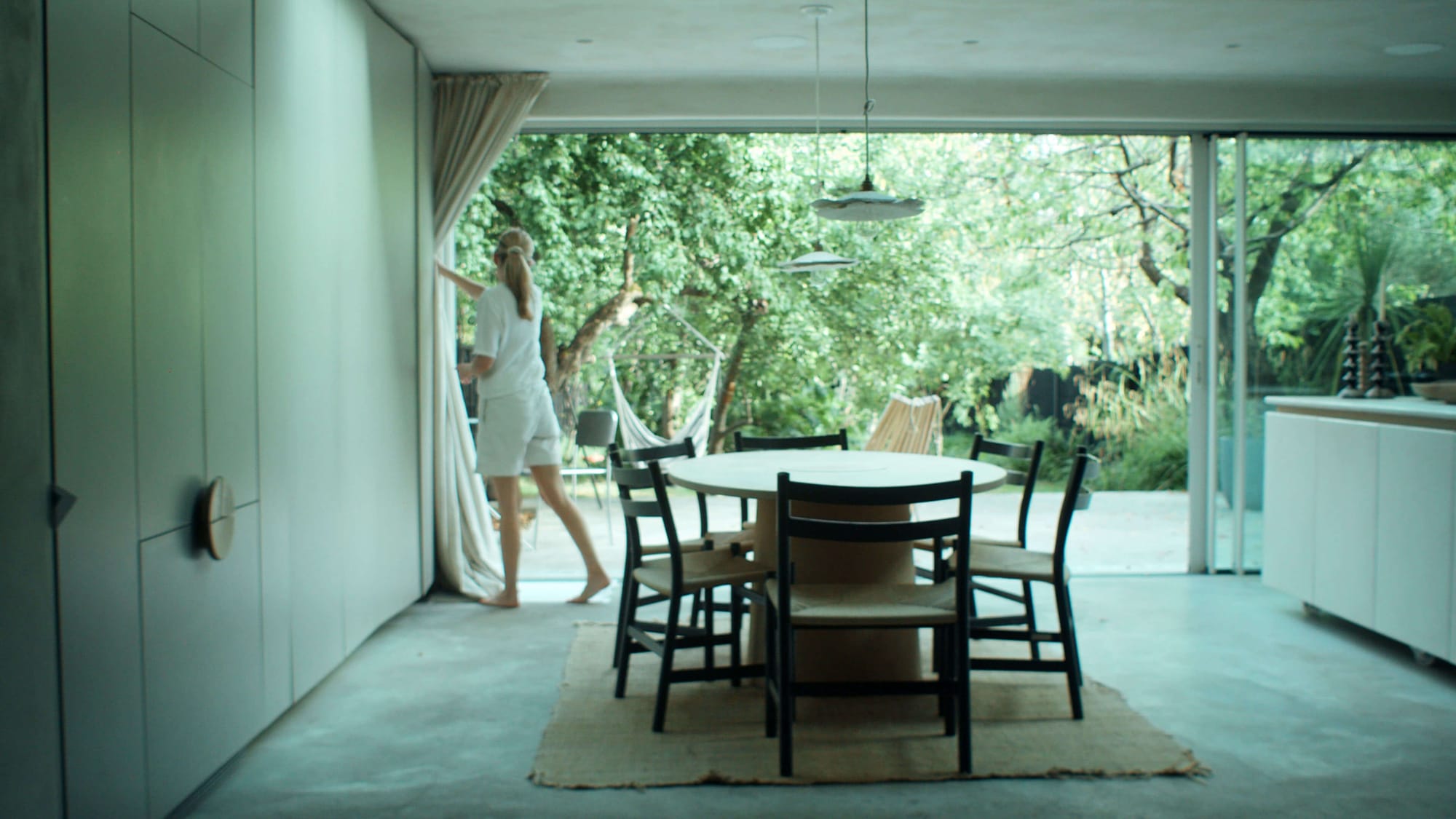Louisa opening curtains in a light-filled dining room with round table, black chairs and garden visible through sliding glass doors