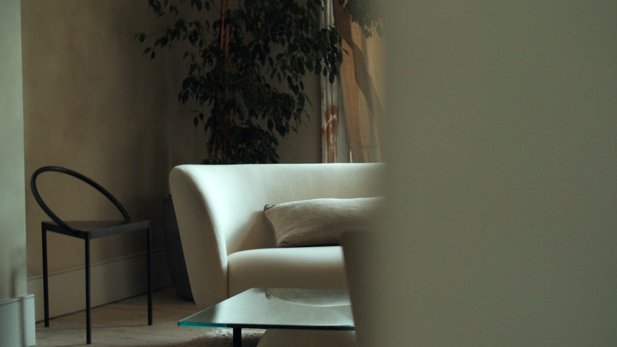 Curved cream sofa with a neutral cushion in a softly lit living room, glass coffee table in the foreground, indoor plant and light curtains in the background