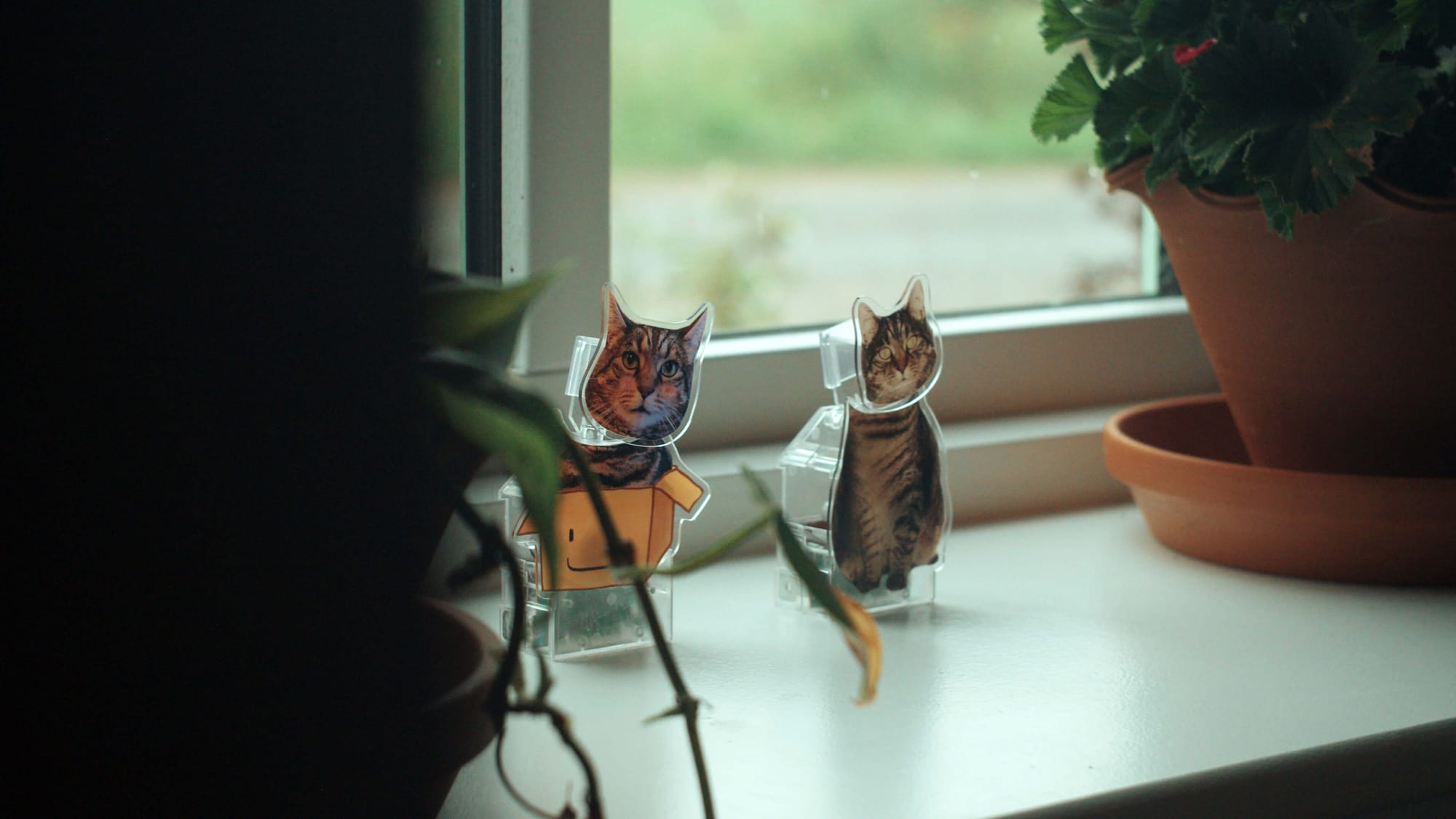 Two small cat figurines on a windowsill beside potted plants