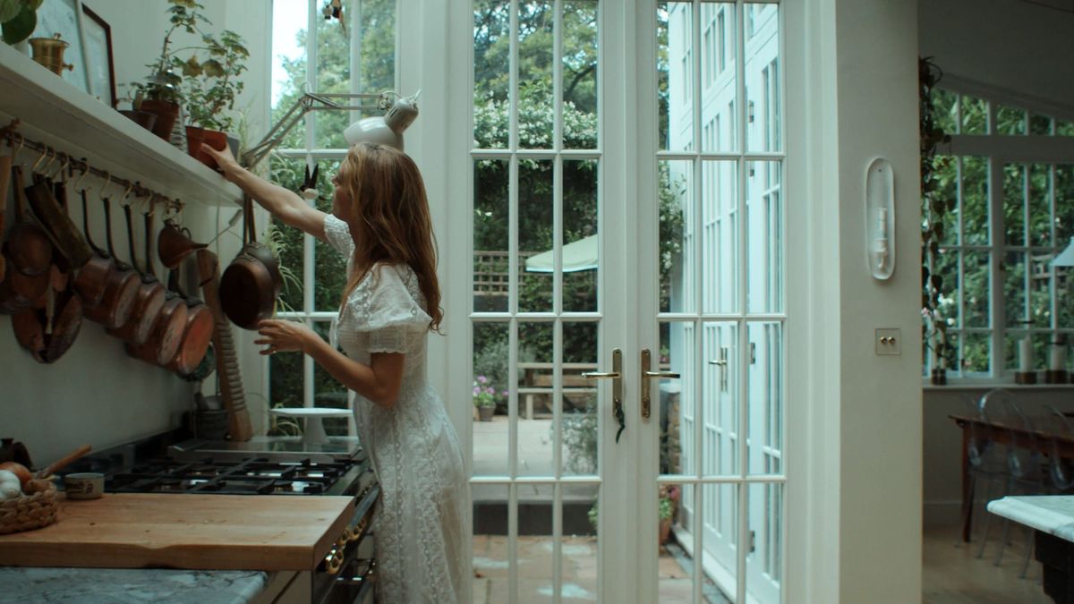 Leanne Kilroy in a white dress reaches for a pot in a bright kitchen with glass doors looking onto a garden