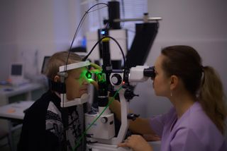An optometrist examines a patient's eyes in a clinical setting using specialized equipment.
