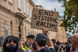 A protester holds a hand-lettered cardboard sign reading 'End Systemic Racism!' above a crowd gathered outside a tan stone building.