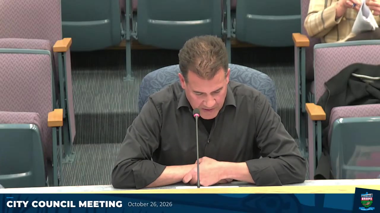 A man in a dark shirt speaks into a microphone at a council table during the Brooks City Council Meeting on October 26, 2026, reading from documents.