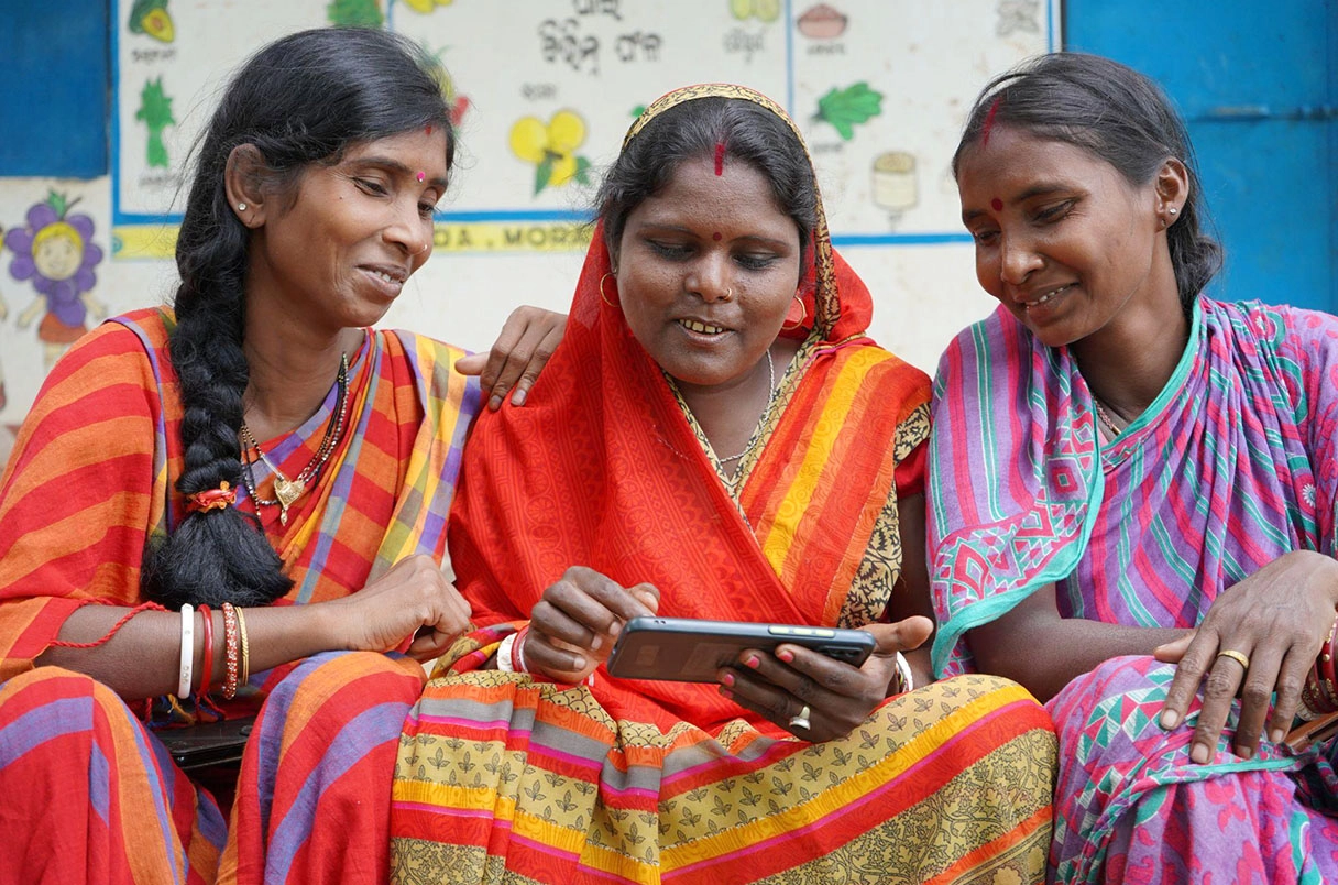 Three Indian women sitting together with their attention focused on something on a phone screen in front of the middle woman.