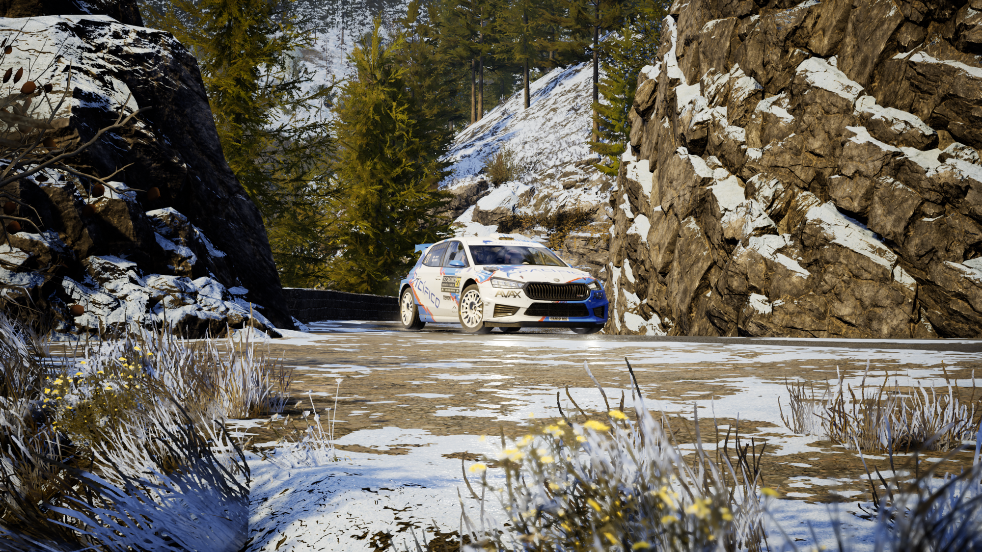 A Skoda Fabia Rally car navigating a turn that's between a rock wall on each side of it. There is plenty of patchy snow on the ground, the rock walls, and the hills behind.