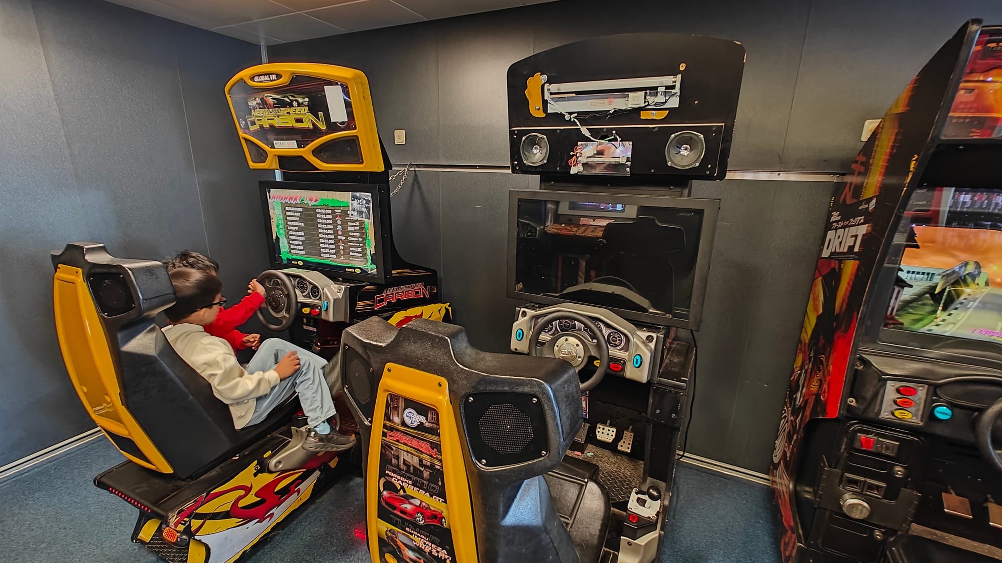 Two kids sharing the seat to a Need For Speed Carbon arcade cabinet on the left, with a broken NFS Carbon arcade cabinet in the middle, and on the right partially out of shot a Fast and The Furious: Drift cabinet is visible. The Need For Speed Carbon cabinet is mostly yellow, with black and white graphics with a red dragon on top of the graphics.