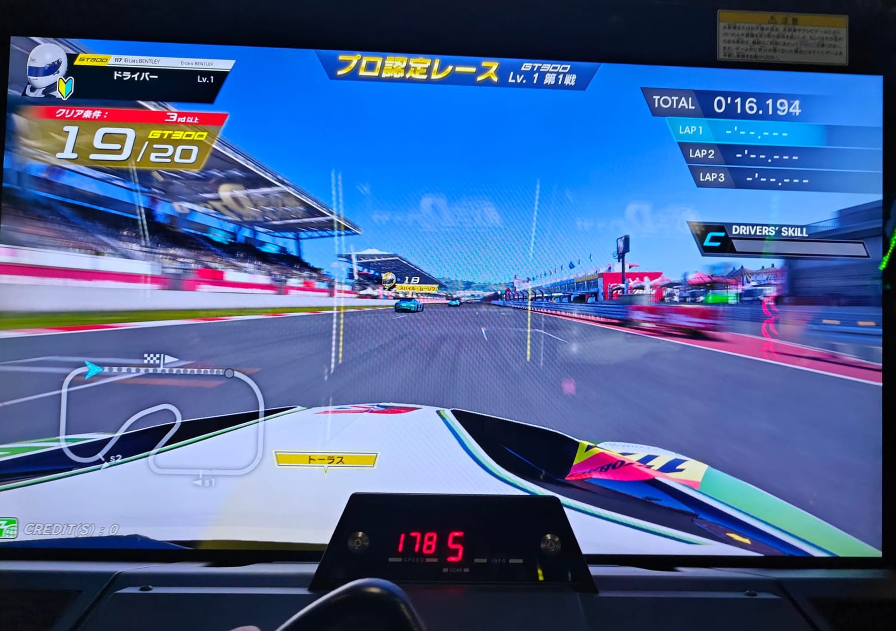 The beginning of a race in SEGA World Drivers Championship featuring Super GT. We are onboard the EI Cars Bentley with it's long white, with light and dark green hood as it's zooms on the front straightaway. The gear and speed indicator are built into the cabinet rather than on the screen, unique for this game.