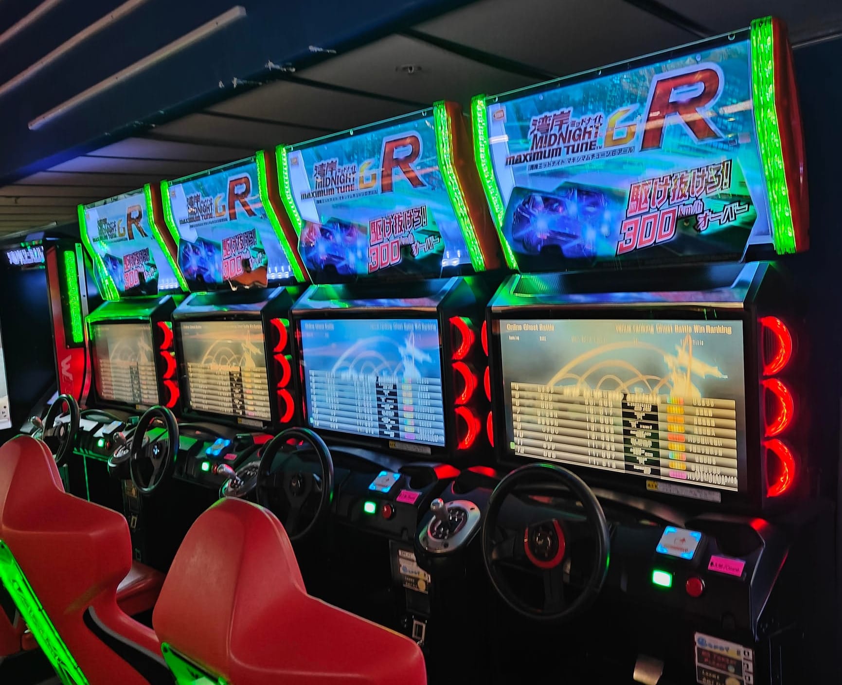 Four Wangan Midnight Maximum Tune 6R arcade racing cabinets in a row. They all have red seats, with the bezels having red and green highlights with the gear shifters being on the left of the steering wheel on each cabinet.
