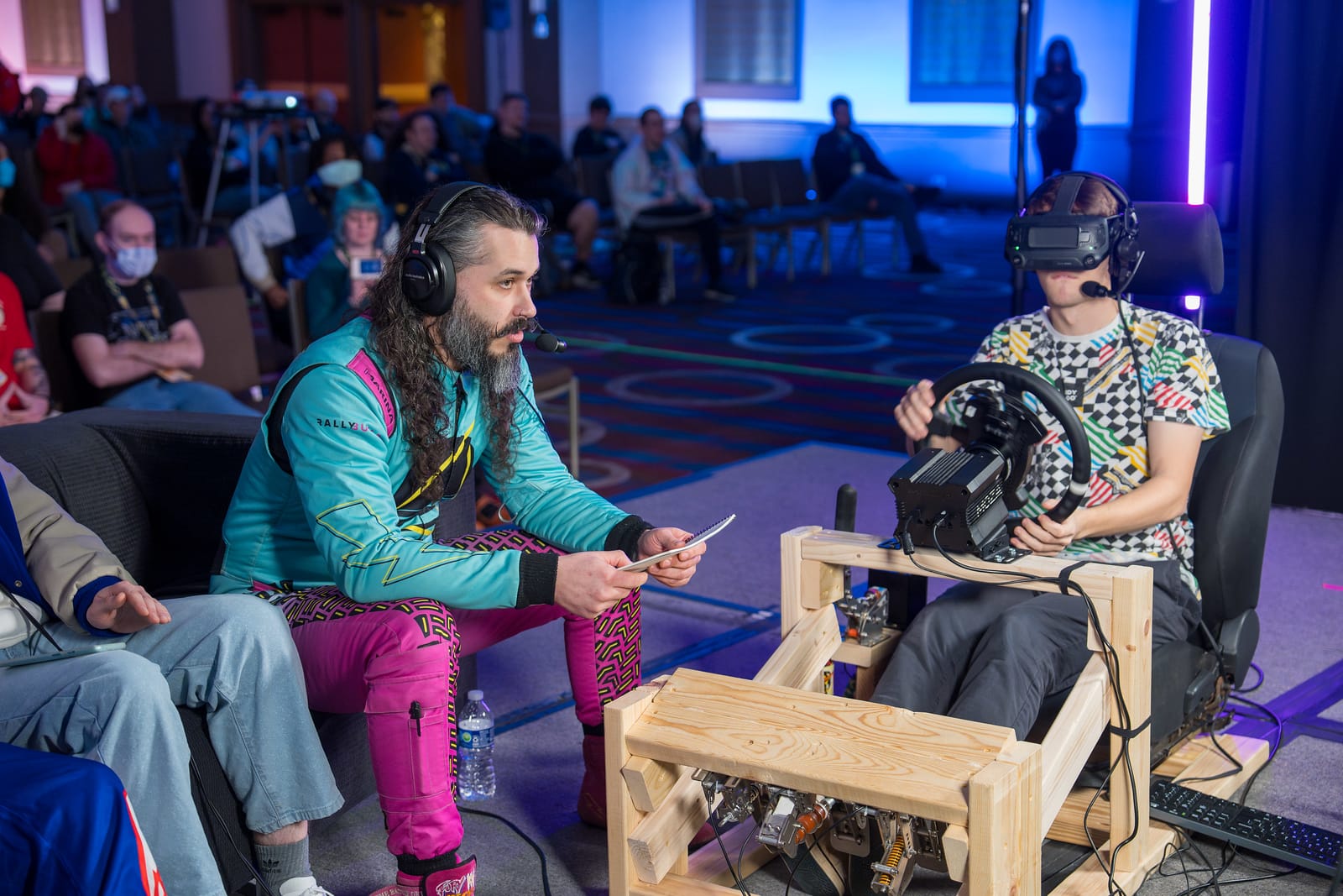 Sisu, wearing the teal and purple drivers suit, is giving live rally pacenotes to Johaan who is driving in Richard Burns Rally on a wooden sim rig while in virtual reality.