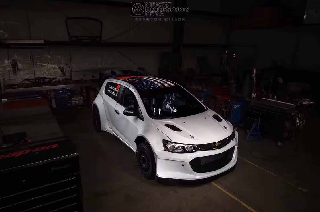 A white Chevy Sonic hatchback with a USA flag graphic on the roof parked in a dimly lit workshop.