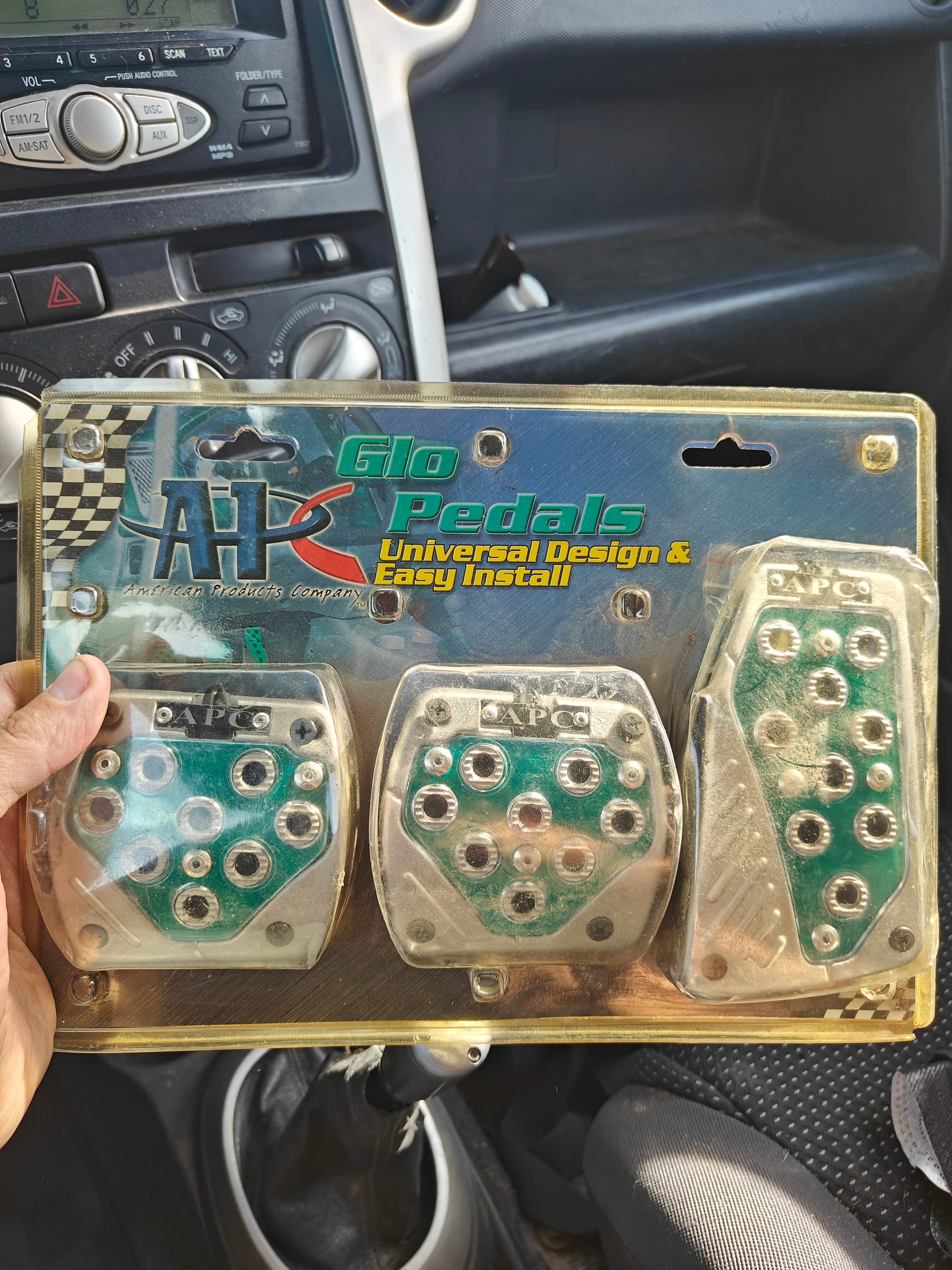 Holding up the APC Glo Pedals while in the car. The blister pack is significantly sun burned and the top corner of the plastic has been chipped away. The pedals inside are silver with a massive green section in the middle and the packaging says "APC Glo Pedals, Universal Design & Easy Install"