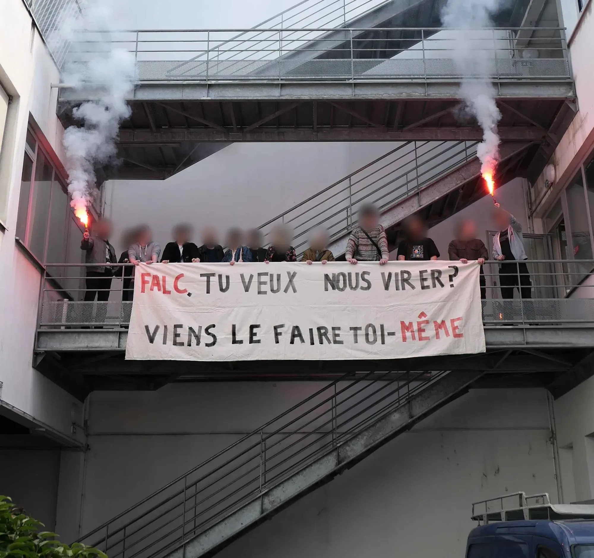 A group of striking workers from Kylotonn picking outside of their studio. The striking workers on the outside of the row, with all of them standing on a mezzanine, have lit flares. They have a banner haning over the handrail that says, "Falc, tu veux nous virer ? Viens le faire toi-même" The banner translated to English says 'Falc (Alain Falc), you want to fire us? Come and do it yourself."