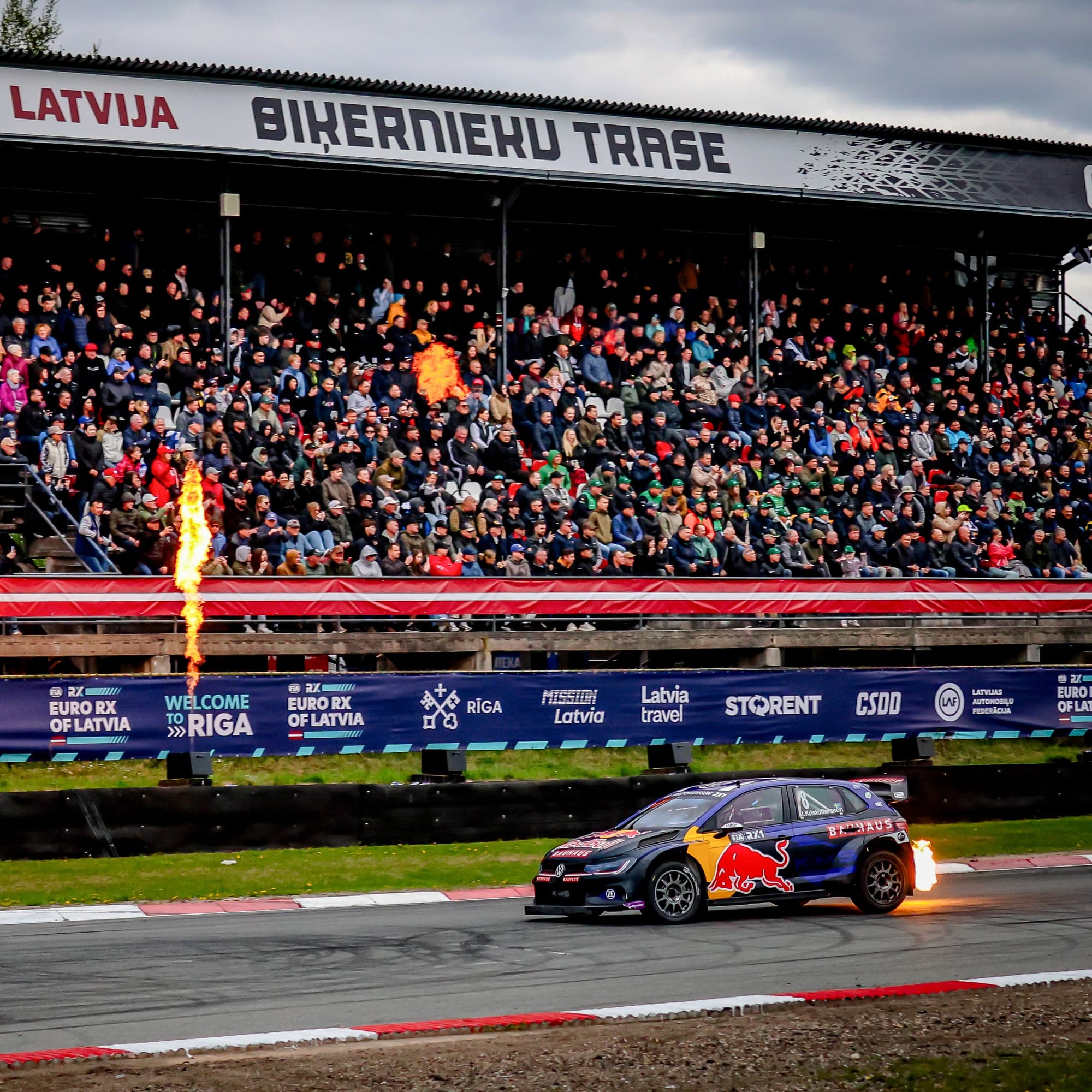 Johan Kristoffersson at the finish of the finals at Biķernieki, Latvia, pyrotechnics fire going off against the barrier with a pack grandstand looking on.