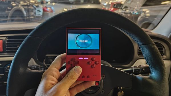 A red Trimui Brick handheld console behind held up against a steering wheel in a car. On the scrren, the intro of Ridge Racer Type 4 is playing.