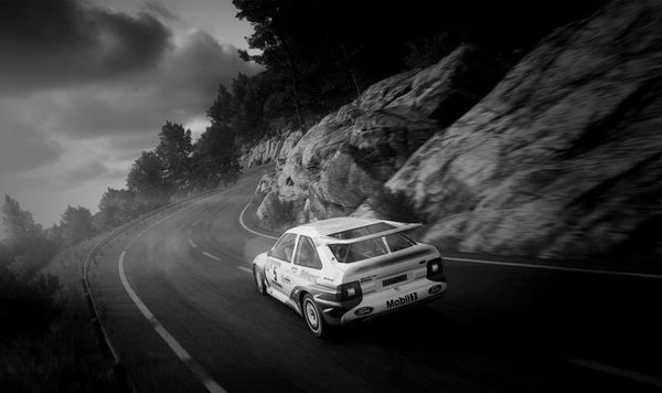 A Ford Escort RS Cosworth rally car on a paved backroad, with boulders to it's right and a sheer cliff guaraded by a armco barrier to it's left in DiRT Rally 2.0