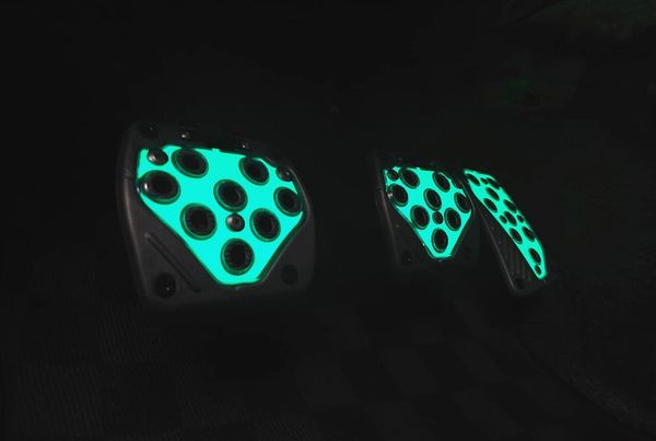 Glowing green pedals in the footwell of a car. There's the longer gas peal on the right side, with the more square brake and clutch pedal alongside.