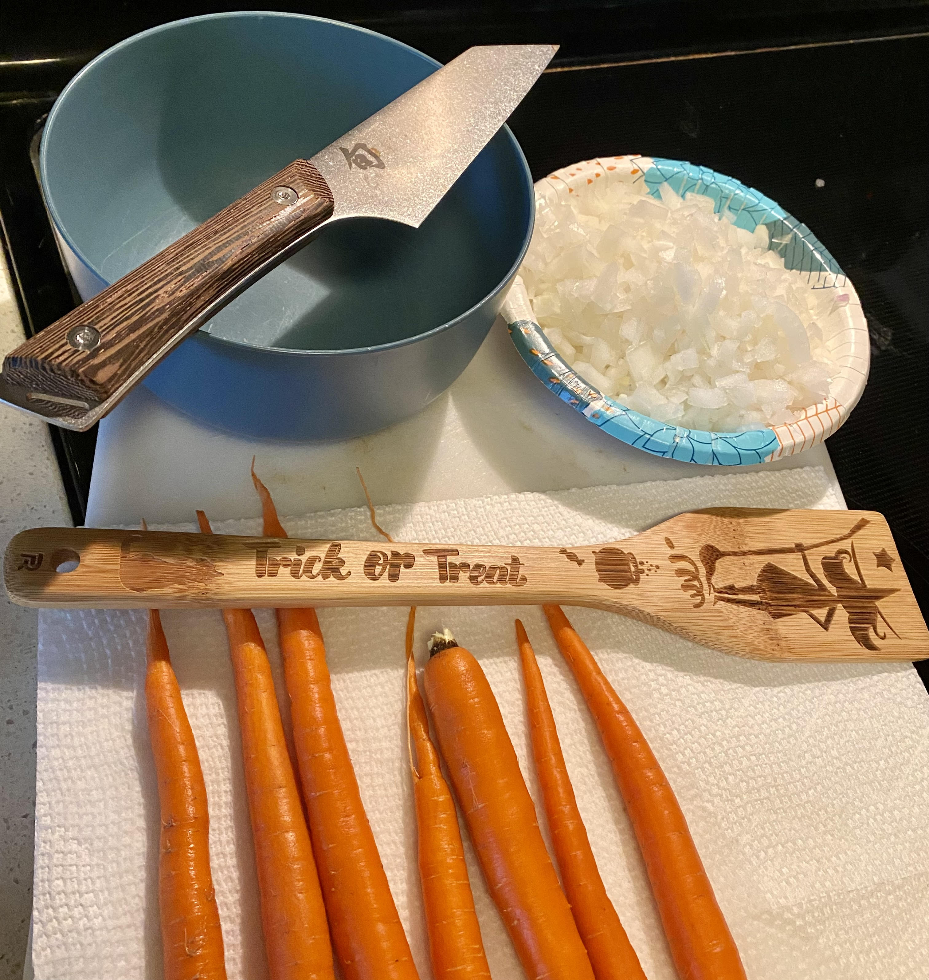 A picture of two bowls, one empty with a sharp knife balanced on top of it, one full of chopped onions, shallots, and garlic, sitting on top of a cutting board. The cutting board is partially covered with paper towel on which some freshly washed carrots have been laid out, and laid across the carrots horizontally is a wooden spatula for sauteeing decorated with an image of a witch, a cat, a cauldron, and the phrase "Trick or Treat."
