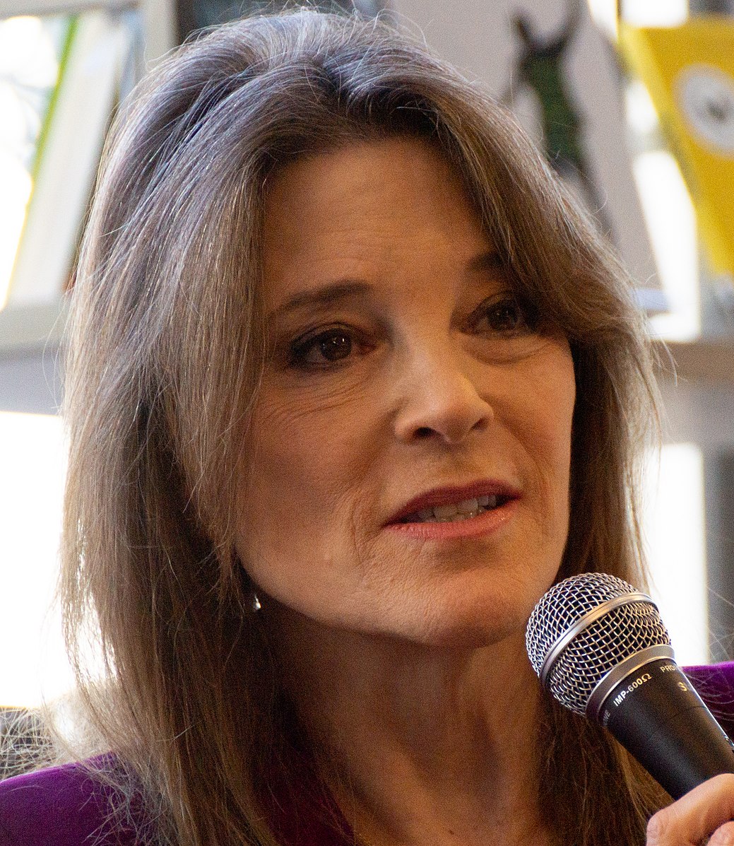 A picture of Marianne Williamson's face--she's a white woman with gray hair and is poised to speak into a microphone.