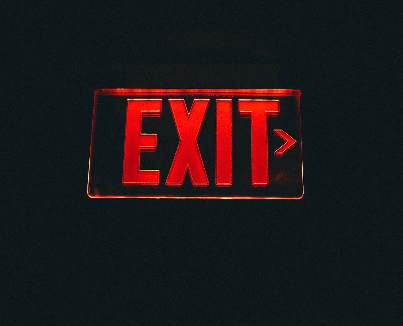 A glass sign lit up with red light illuminating the letters "E-X-I-T" on a dark black background. It almost looks as if the exit sign is imply floating in a black void.