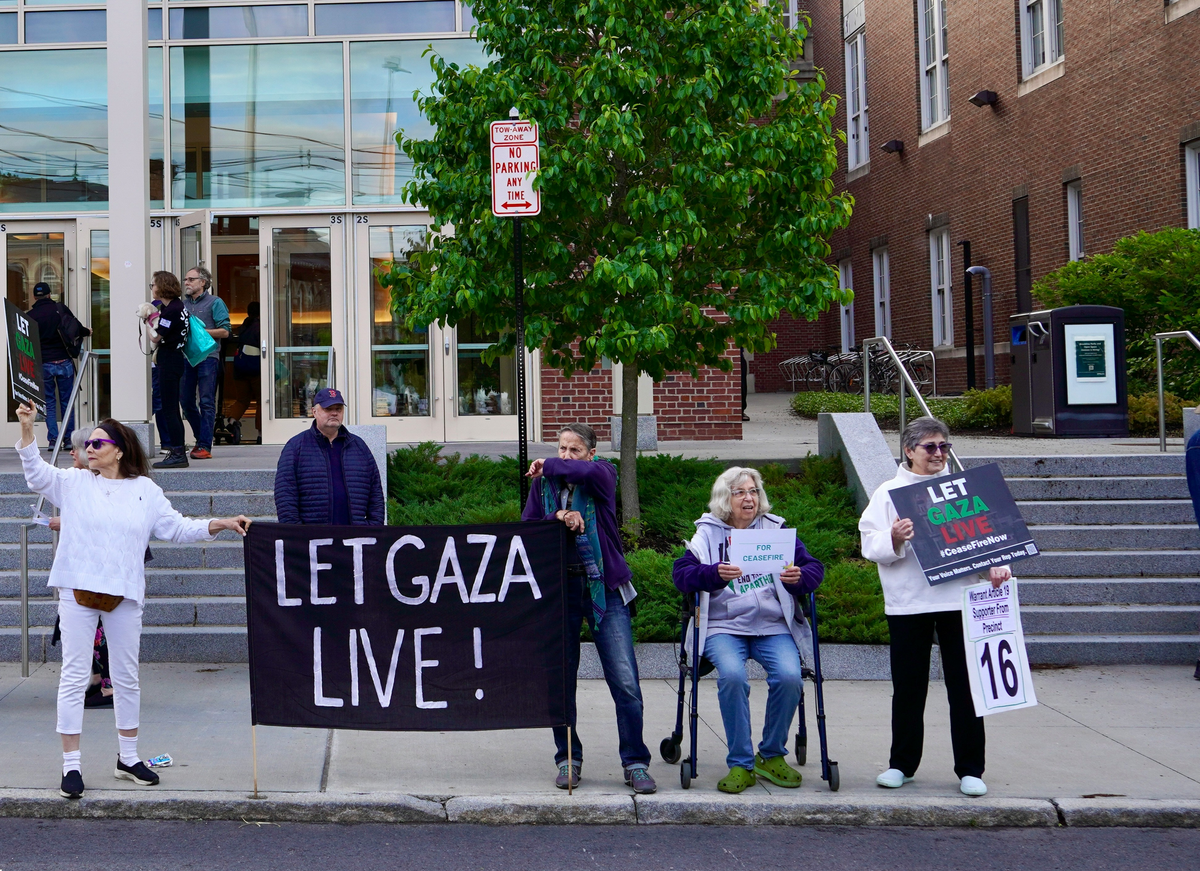 Protesters, most standing and one seated, standing on a sidewalk. Two are holding a large black banner with "LET GAZA LIVE!" on it in large block letters.