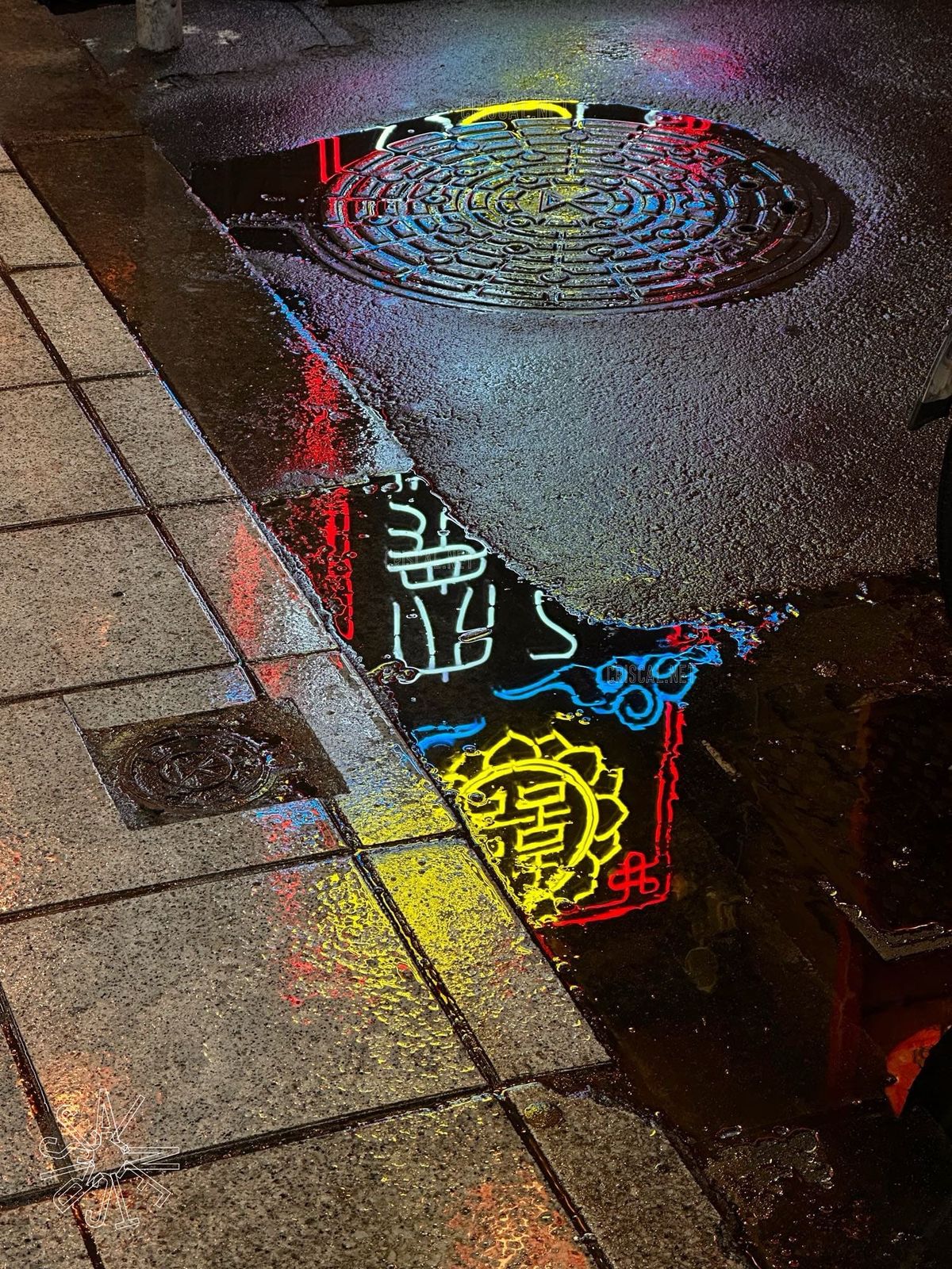 Wet pavement reflecting colorful neon signs in red, yellow, and blue beside a manhole cover, creating a glowing scene at night.