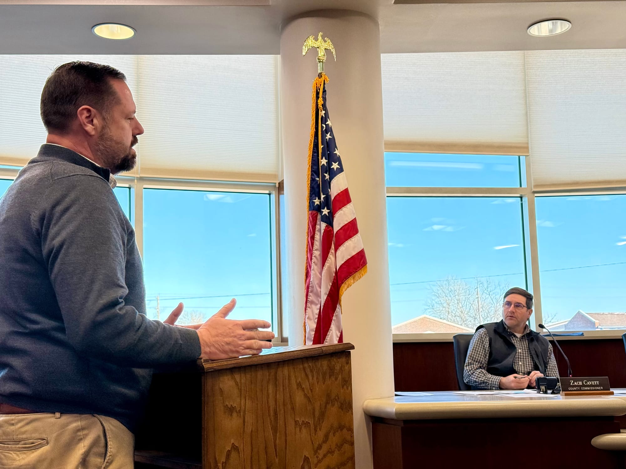 Joyce stands at lectern with Cavett sitting behind the horseshoe table.