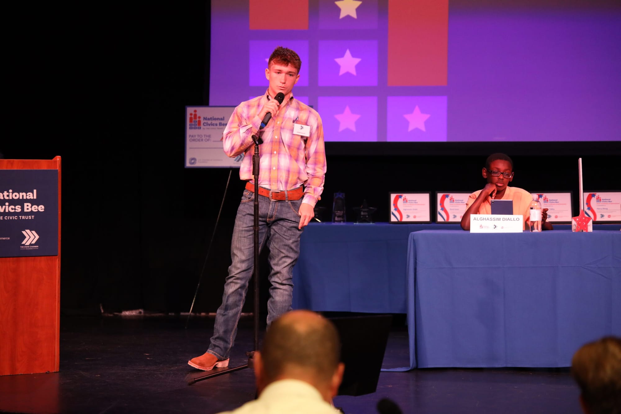 Stran Talley holding a microphone on stage wearing a plaid button shirts, jeans and boots.