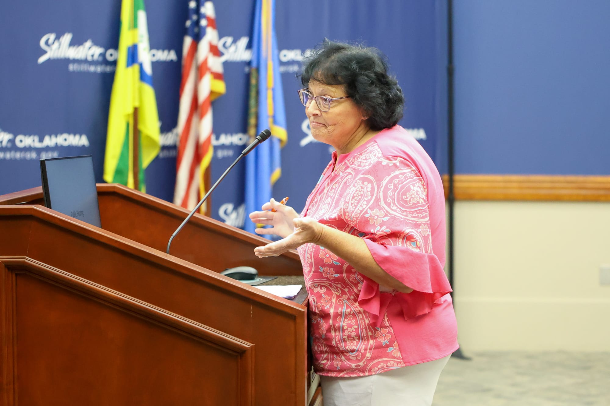 Crystal Hayes gestures while speaking at a podium before the Stillwater Planning Commission, wearing a pink paisley blouse, with American, Oklahoma, and city flags in the background.