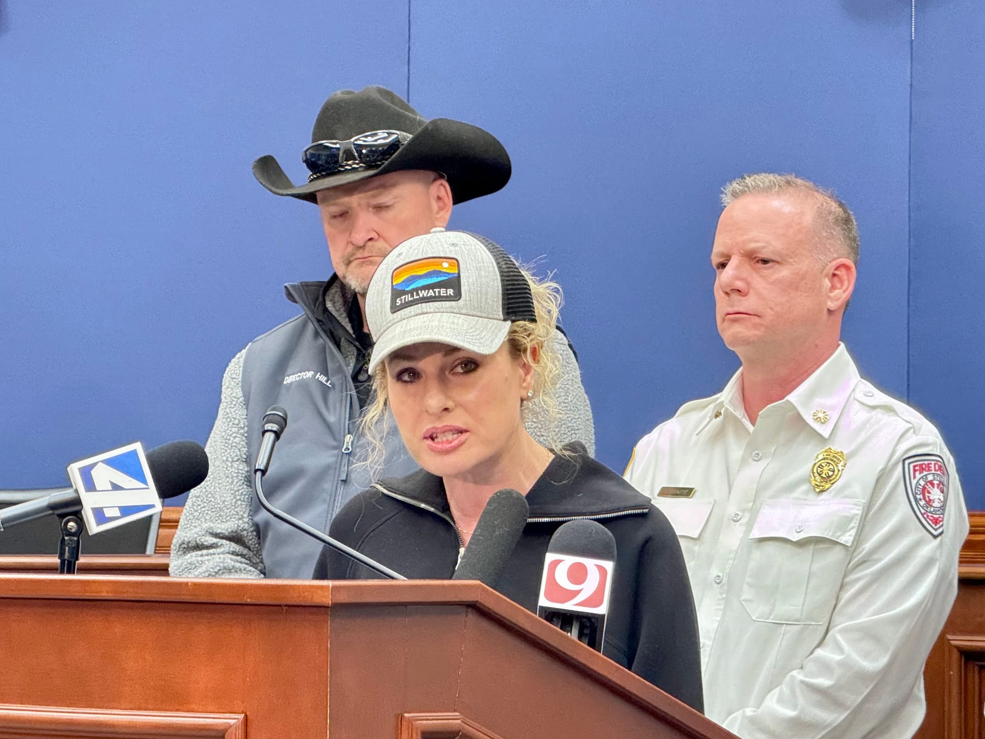 Woman in baseball cap speaks at podium with microphones, flanked by two officials including one in fire department uniform.