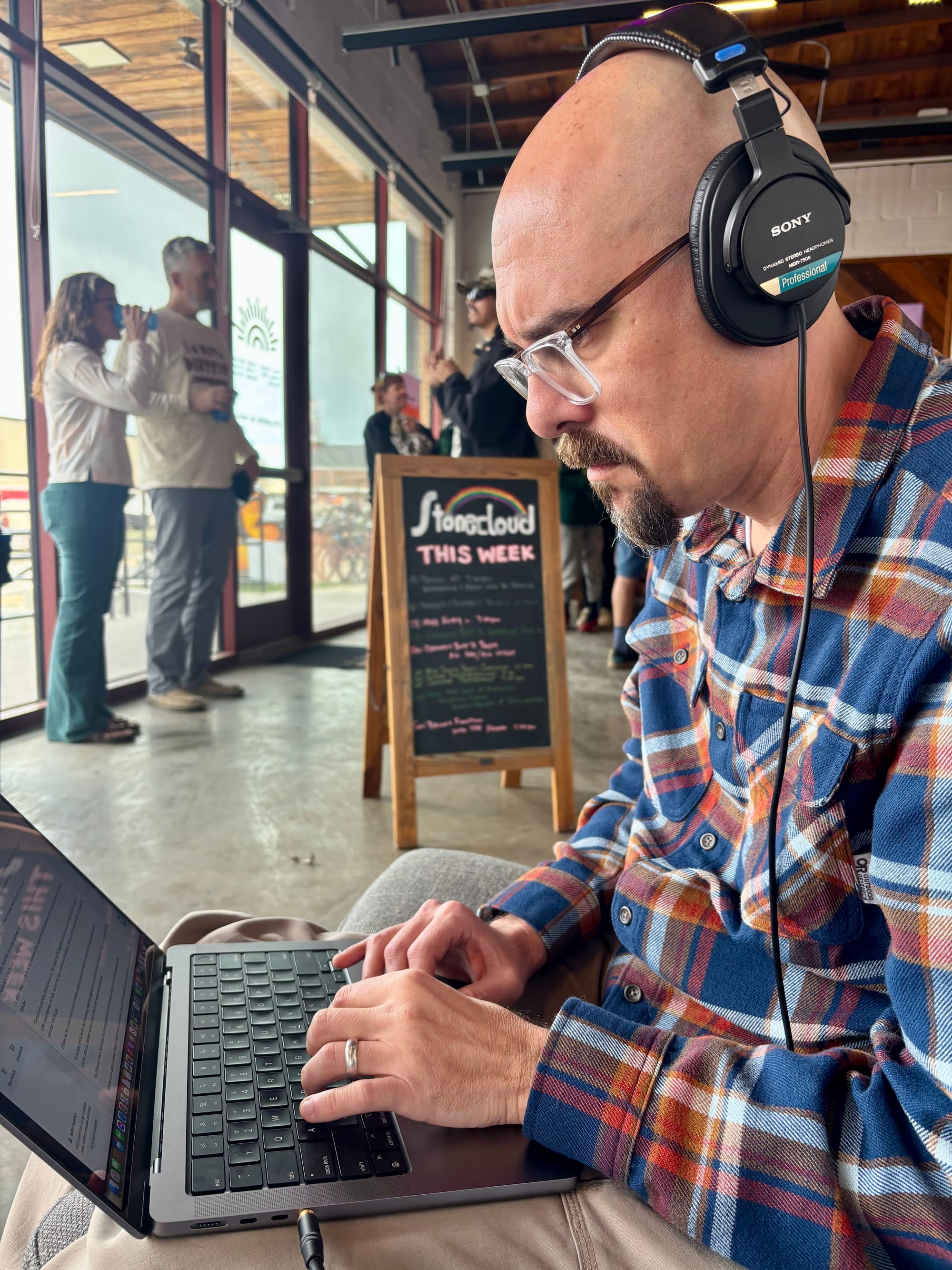 Chris Peters wears headphones and works on a laptop at a table inside Stonecloud Brewery. He's focused on the screen while people gather and talk in the background. A brewery chalkboard sign is visible behind him in the industrial-style space with large windows.