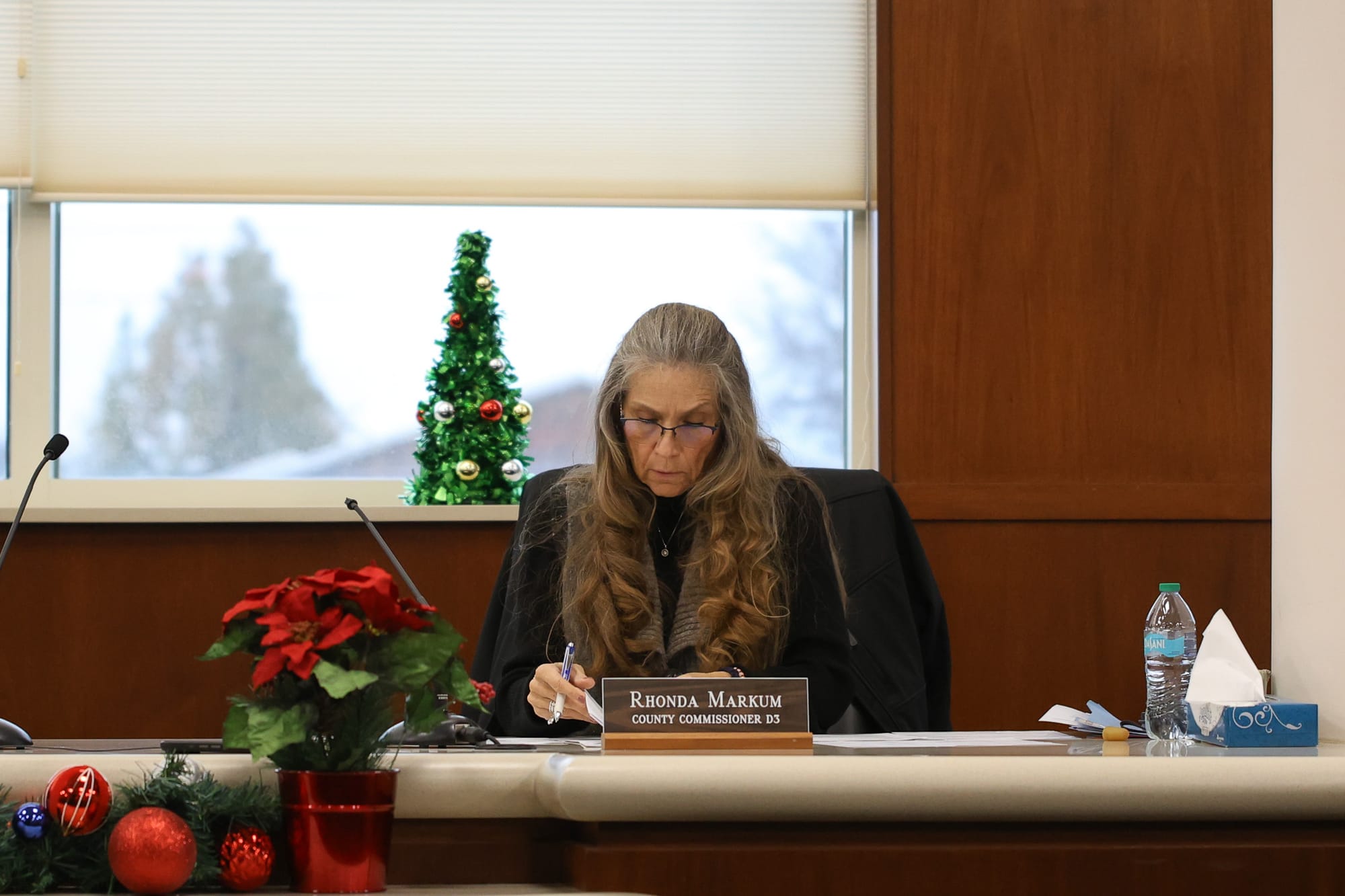 Payne County Commissioner Rhonda Markum sits at the commissioners' bench writing notes, wearing dark clothing and glasses, with her nameplate visible. A small Christmas tree and poinsettia decorate the room.