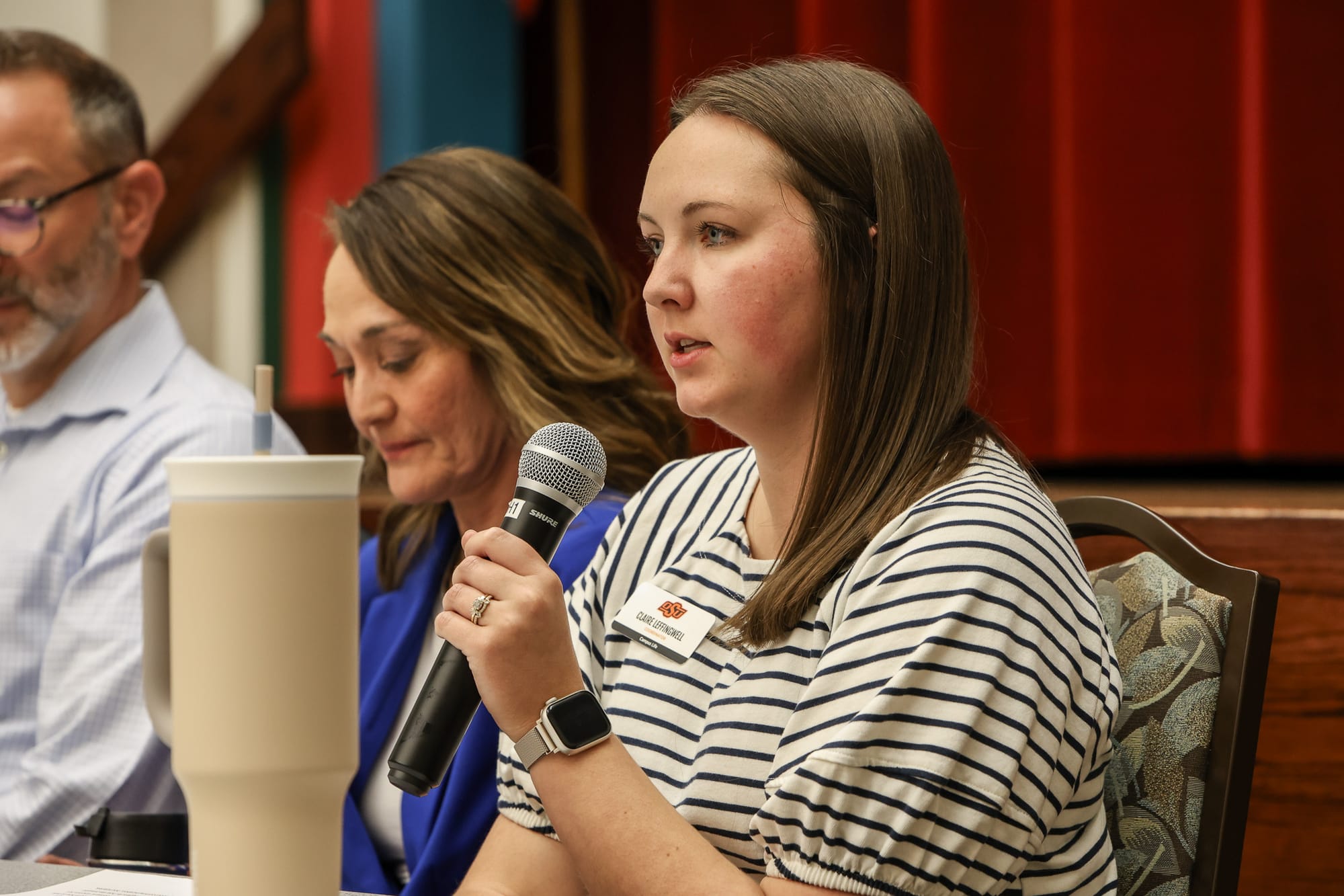 Woman in striped shirt speaks into microphone at panel discussion table, with fellow panelists seated nearby and red curtains visible in the background.