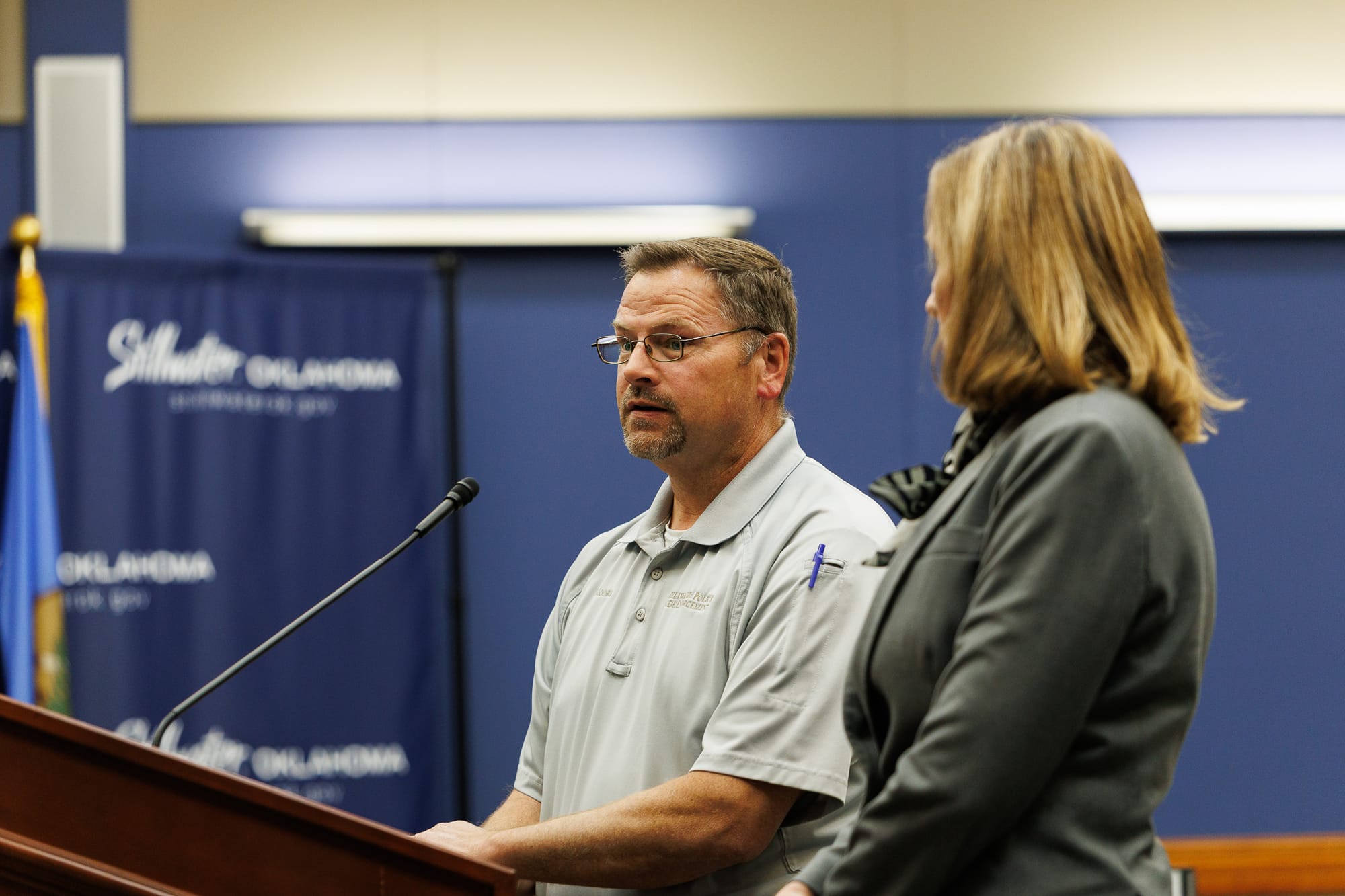 Man in gray polo shirt and glasses speaking at podium with microphone during City Council meeting, with woman in gray blazer standing beside him and blue backdrop visible behind them