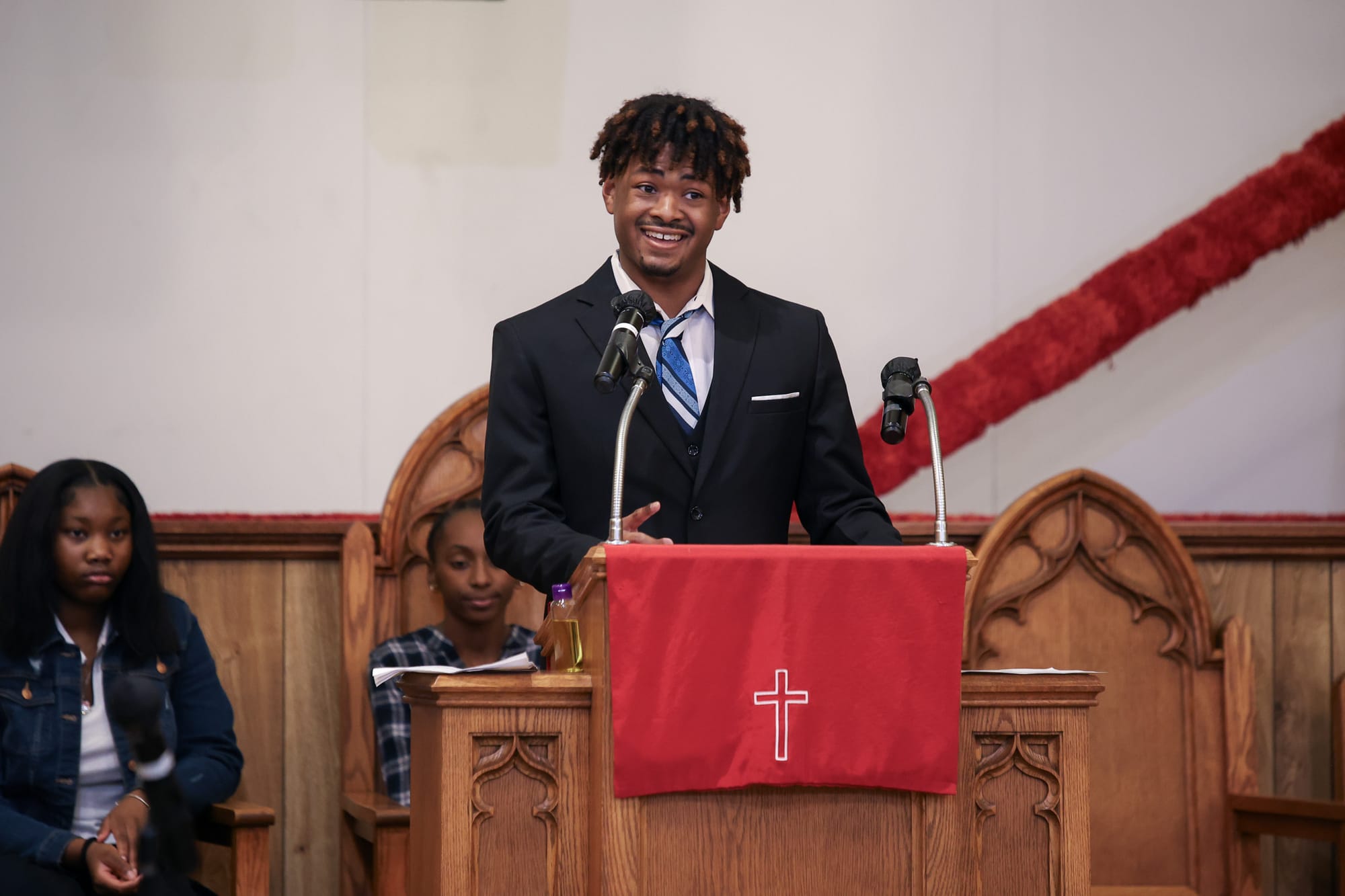 Jamiere Dickens-Smith speaks at podium during MLK Jr. Day celebration at Mt. Zion Baptist Church, wearing black suit and blue striped tie