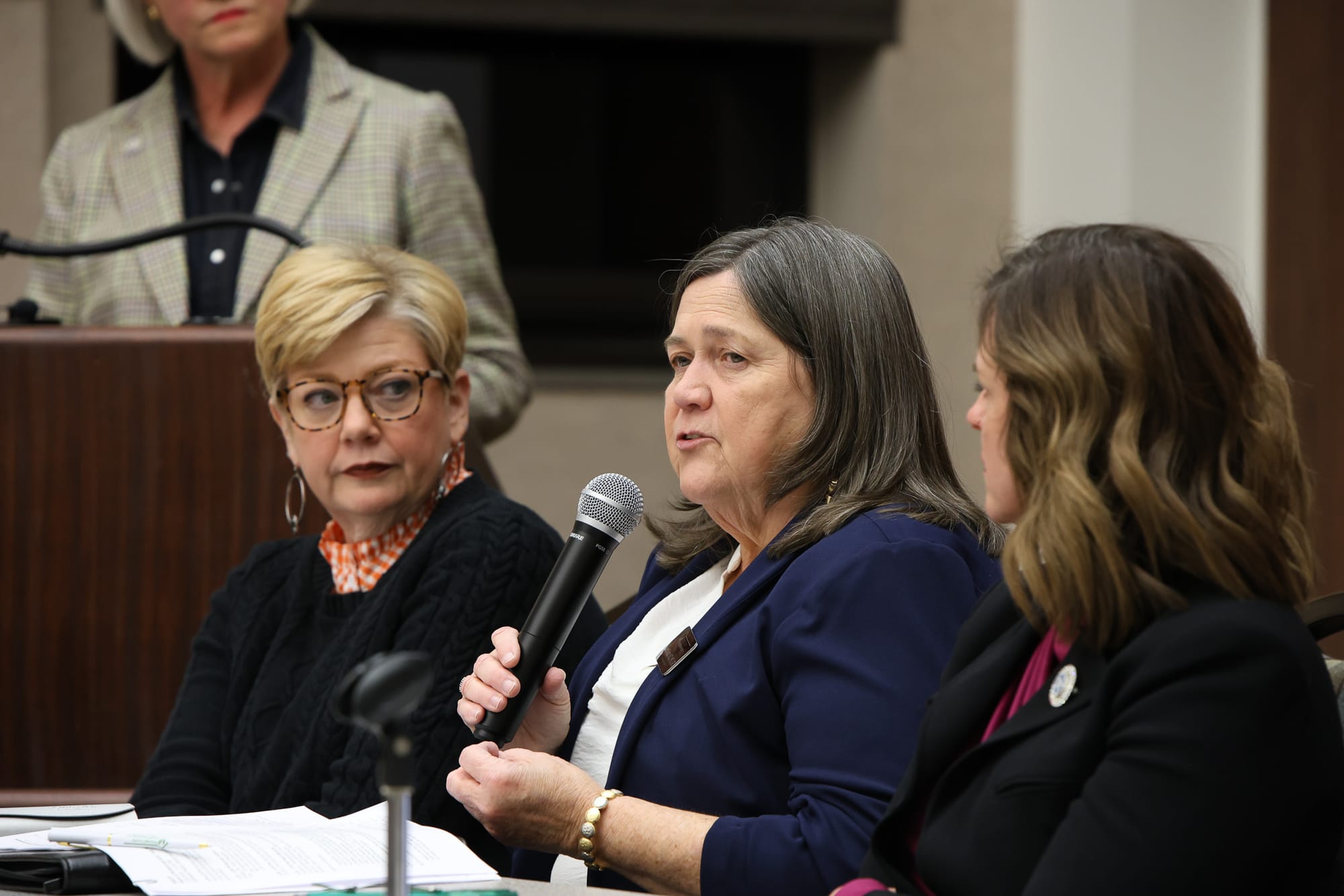 Janet Fultz holds a microphone while speaking at a panel discussion, seated between fellow panelists with a moderator standing at podium in the background.
