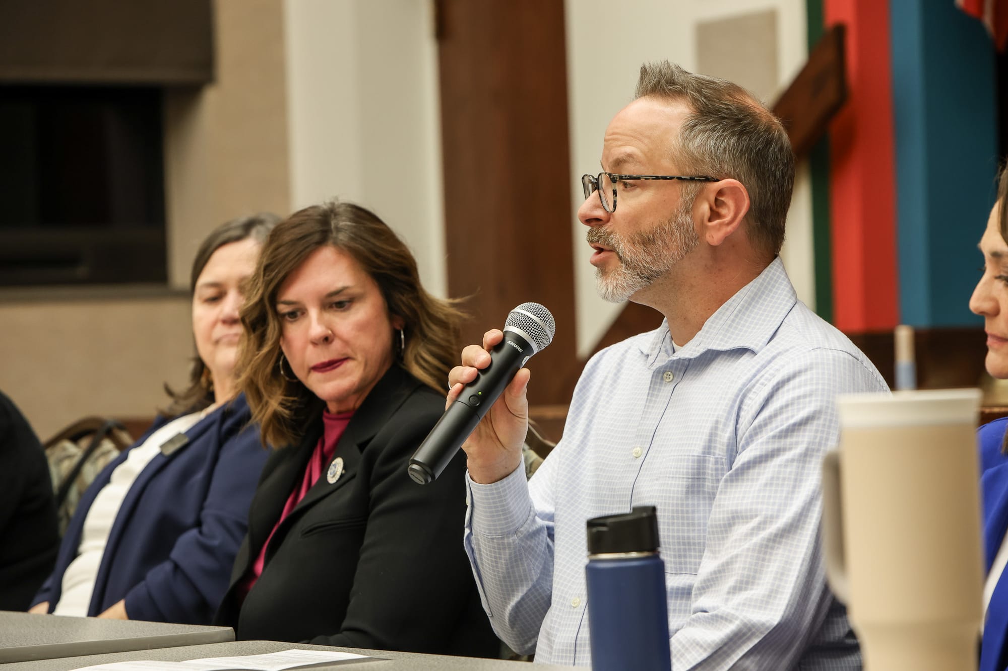 Jimmy Widdifield speaks into a microphone during a panel discussion at Stillwater Public Library, wearing glasses and a light blue checkered shirt, with fellow panelists visible.
