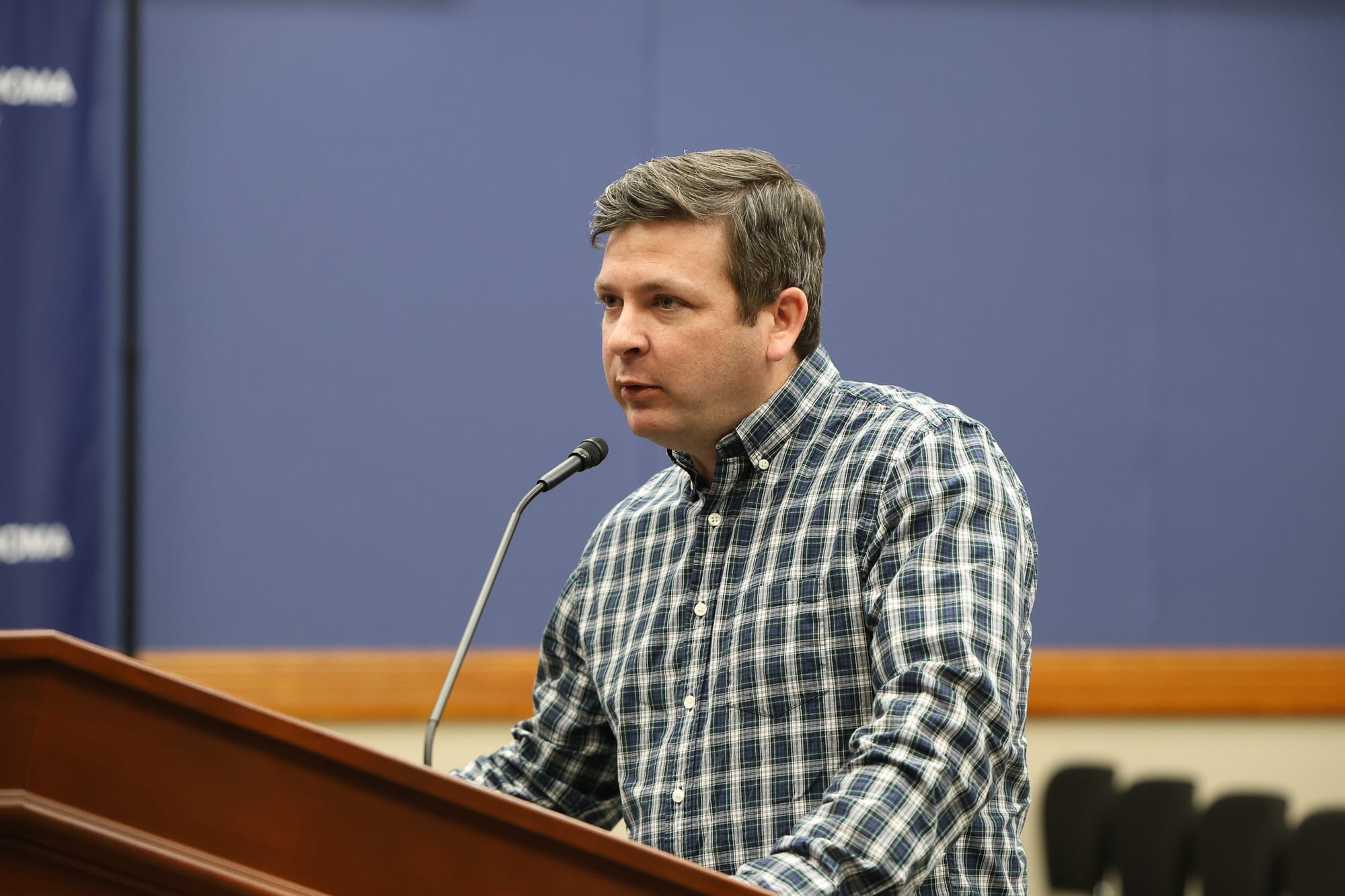 Man in plaid button-down shirt speaking at microphone podium in council chambers with blue wall behind him.