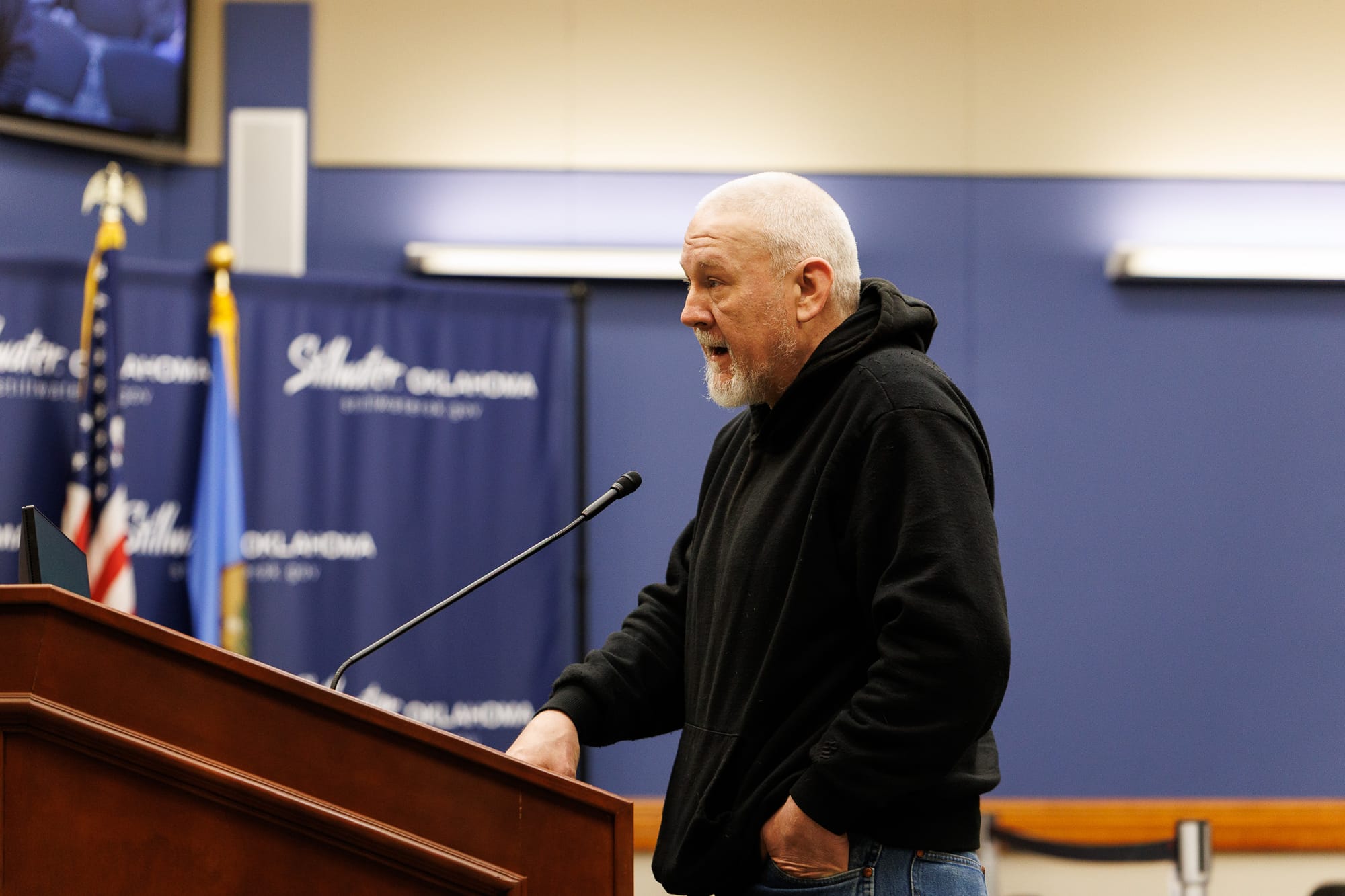 Man with white goatee wearing black hoodie speaking at wooden podium with microphone during City Council meeting, with American flag visible in the background