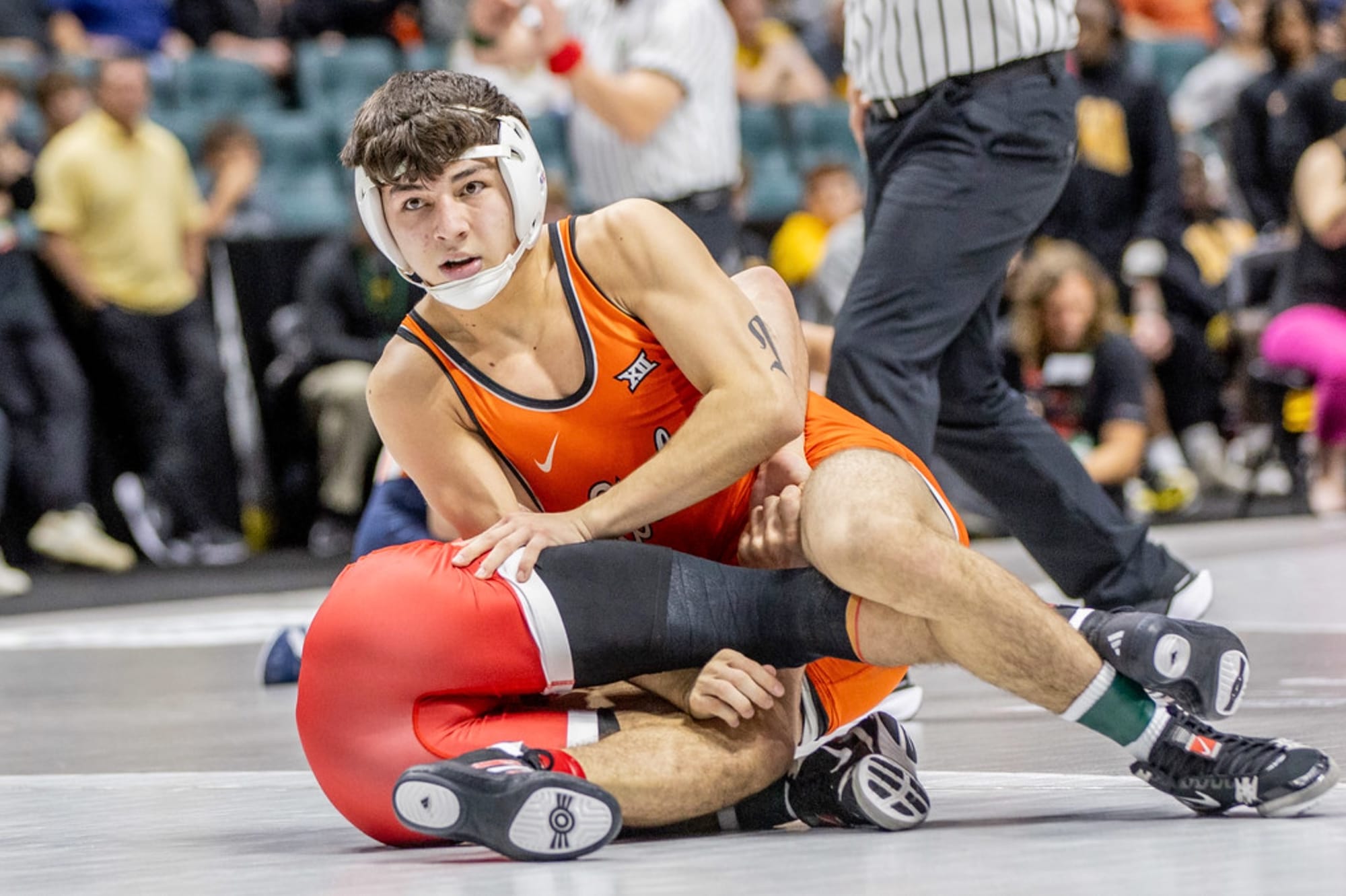 OSU freshman wrestler Ronnie Ramirez in orange singlet competes on the mat during a match against NC State at BOK Center in Tulsa. Ramirez controls position against opponent in red singlet.