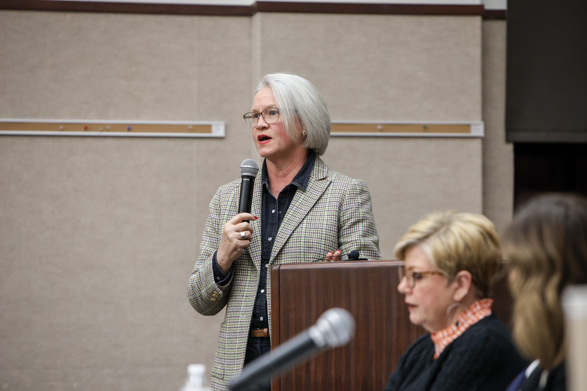 Trish Ranson addresses attendees from podium during panel discussion, wearing patterned blazer and glasses, with panelists visible in the foreground.