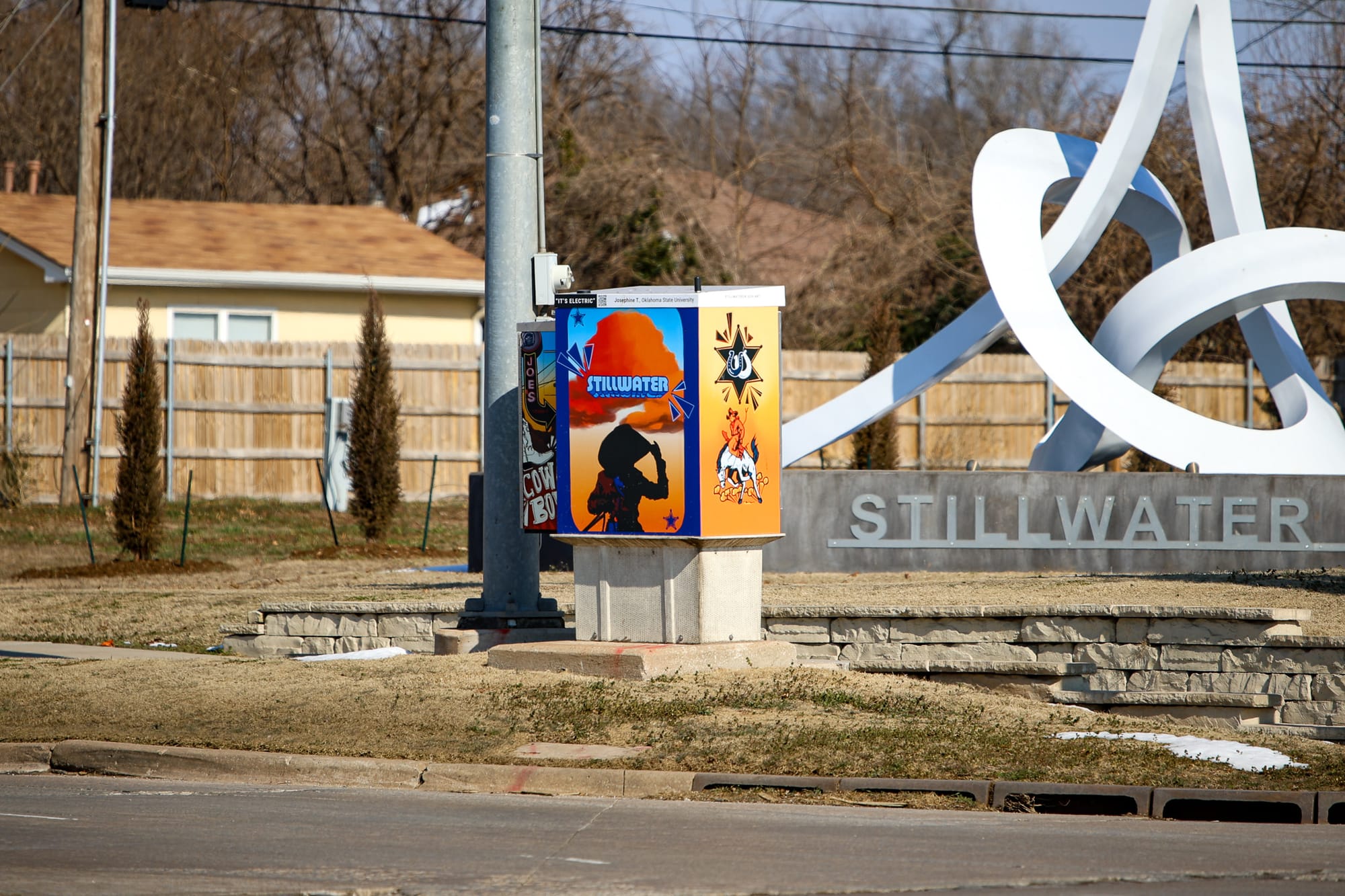 Decorated utility box with colorful artwork showing cowboy silhouette and Stillwater text stands in foreground with white Interlace sculpture and stone wall visible behind it at intersection.