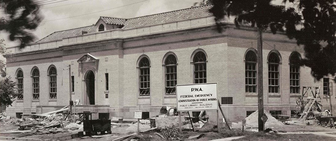 Black and white photo of neoclassical brick library building under construction in 1937, showing arched windows and a PWA Federal Emergency Administration sign in front of the construction site.
