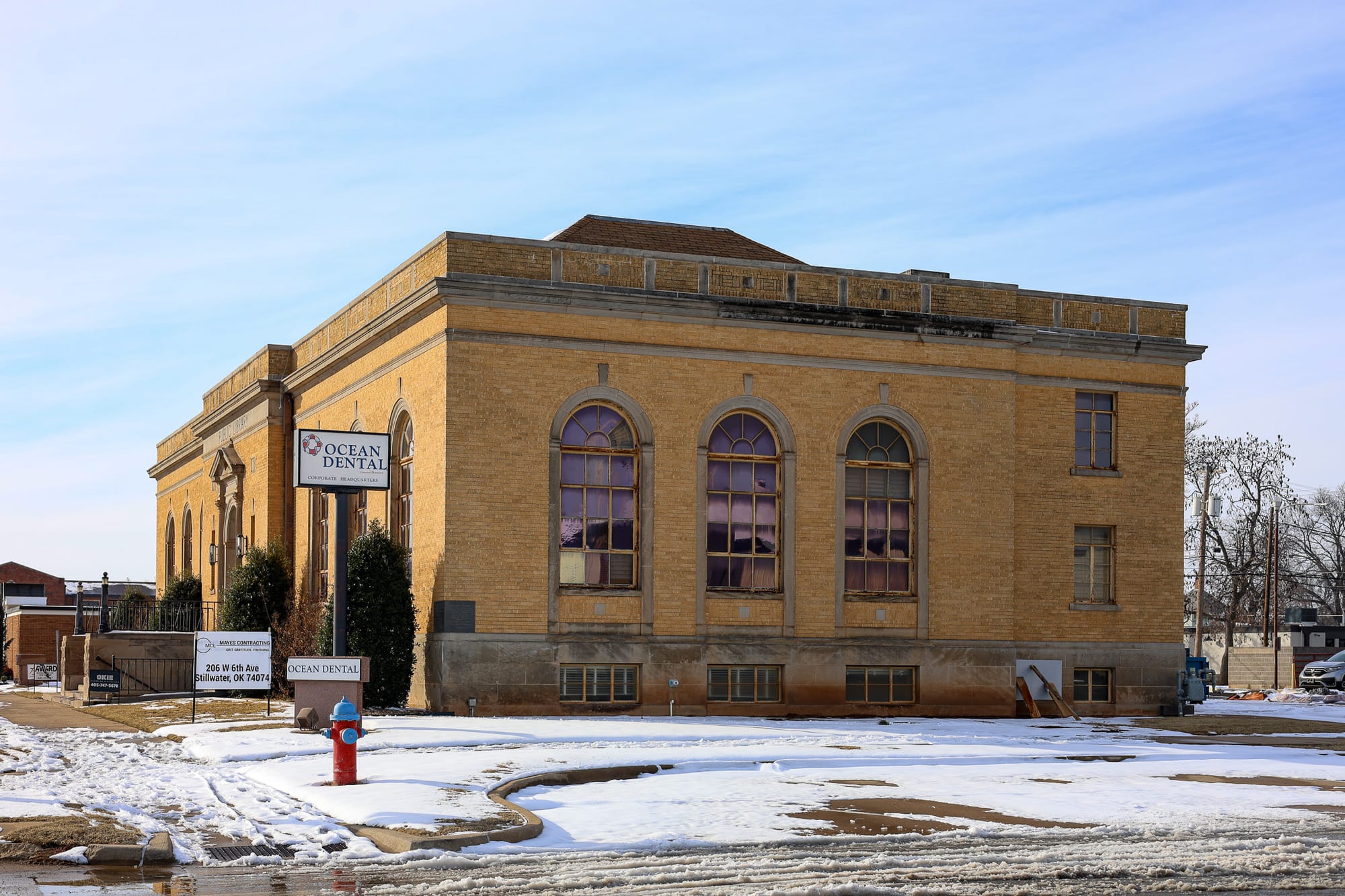 Historic tan brick library building with three large arched windows on the front facade, photographed in winter with snow on the ground and Ocean Dental signage visible.