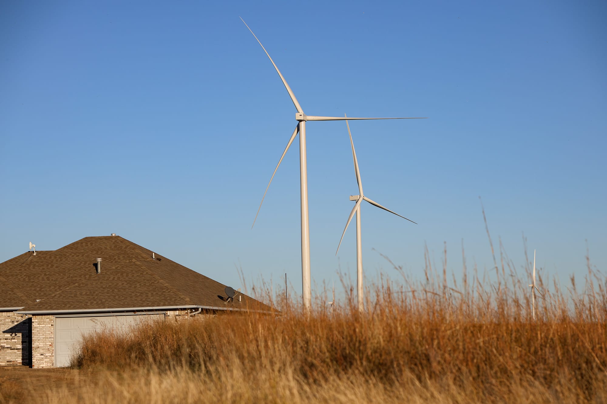 Wind turbines rise behind a brick home with a dark roof, with tall dry prairie grass in the foreground under a clear blue sky in Noble County, Oklahoma