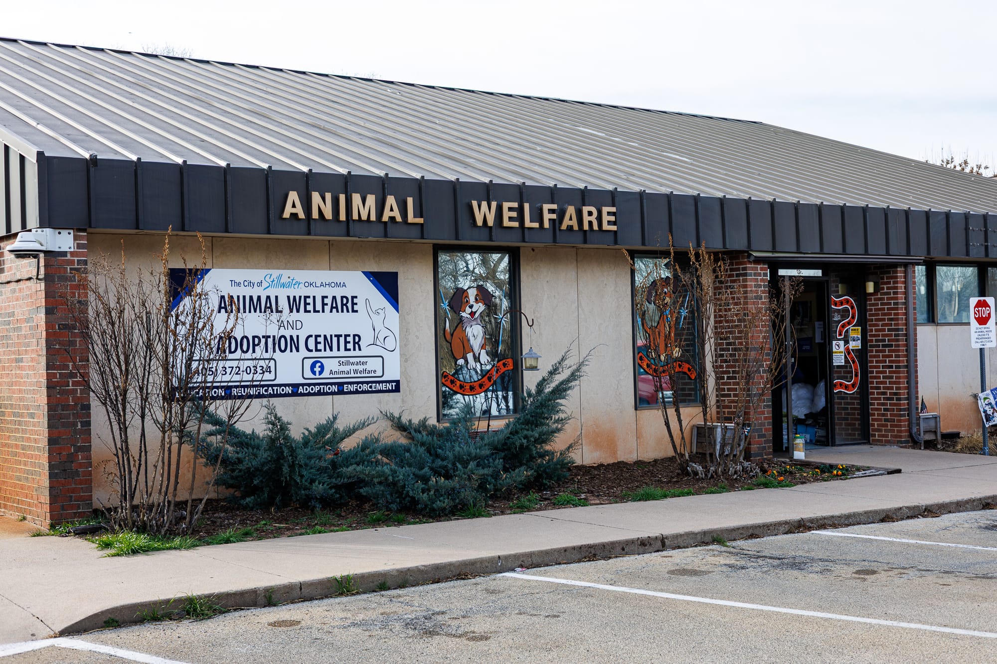 Stillwater Animal Welfare building with brick facade, metal roof, "ANIMAL WELFARE" sign above entrance, painted murals on windows, landscaping with bushes along sidewalk