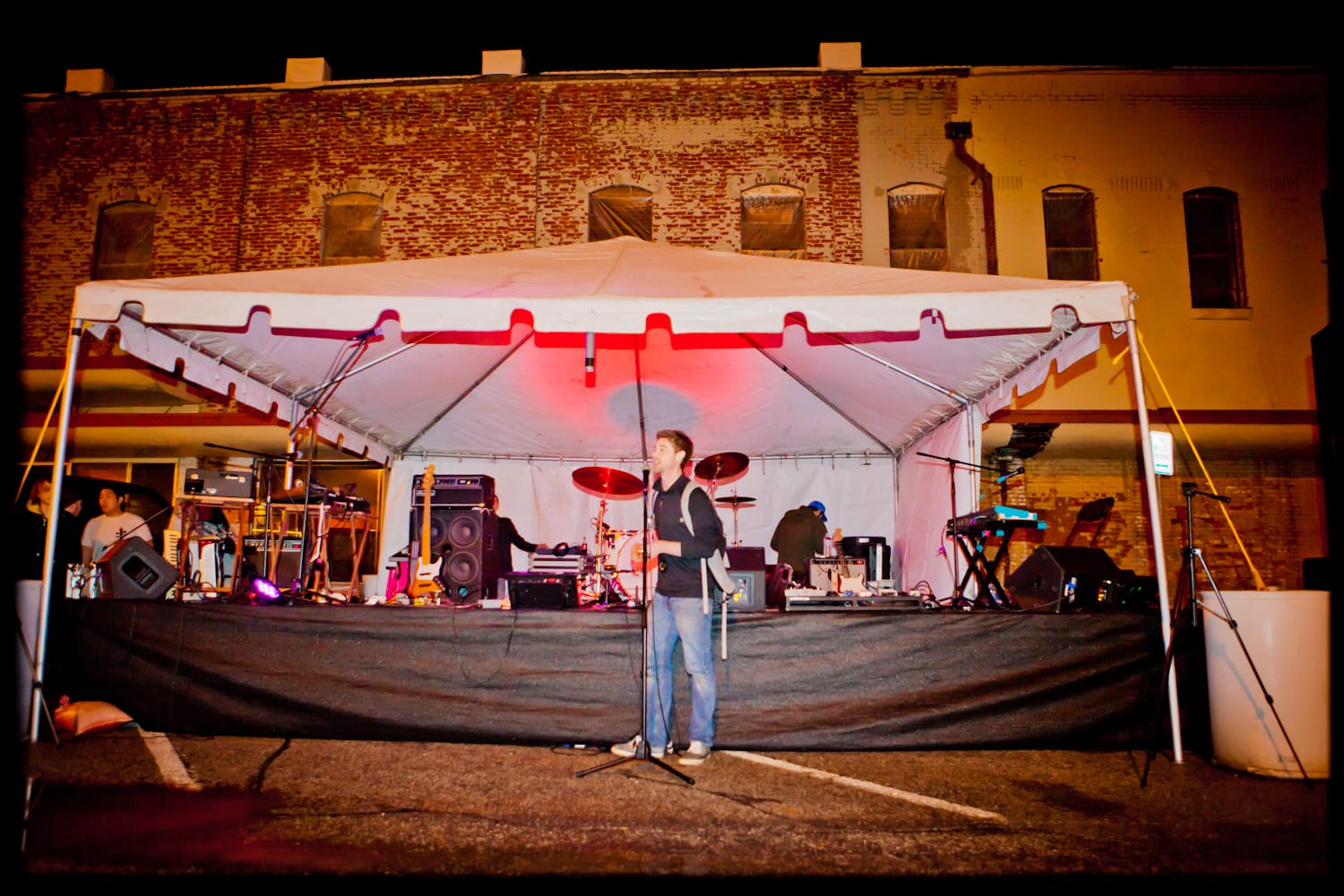 Bobby Wintle stands at a microphone in front of an outdoor tent stage at night while band members set up equipment behind him, with a brick building visible in the background, during the 2013 Land Run 100 in downtown Stillwater, Oklahoma.