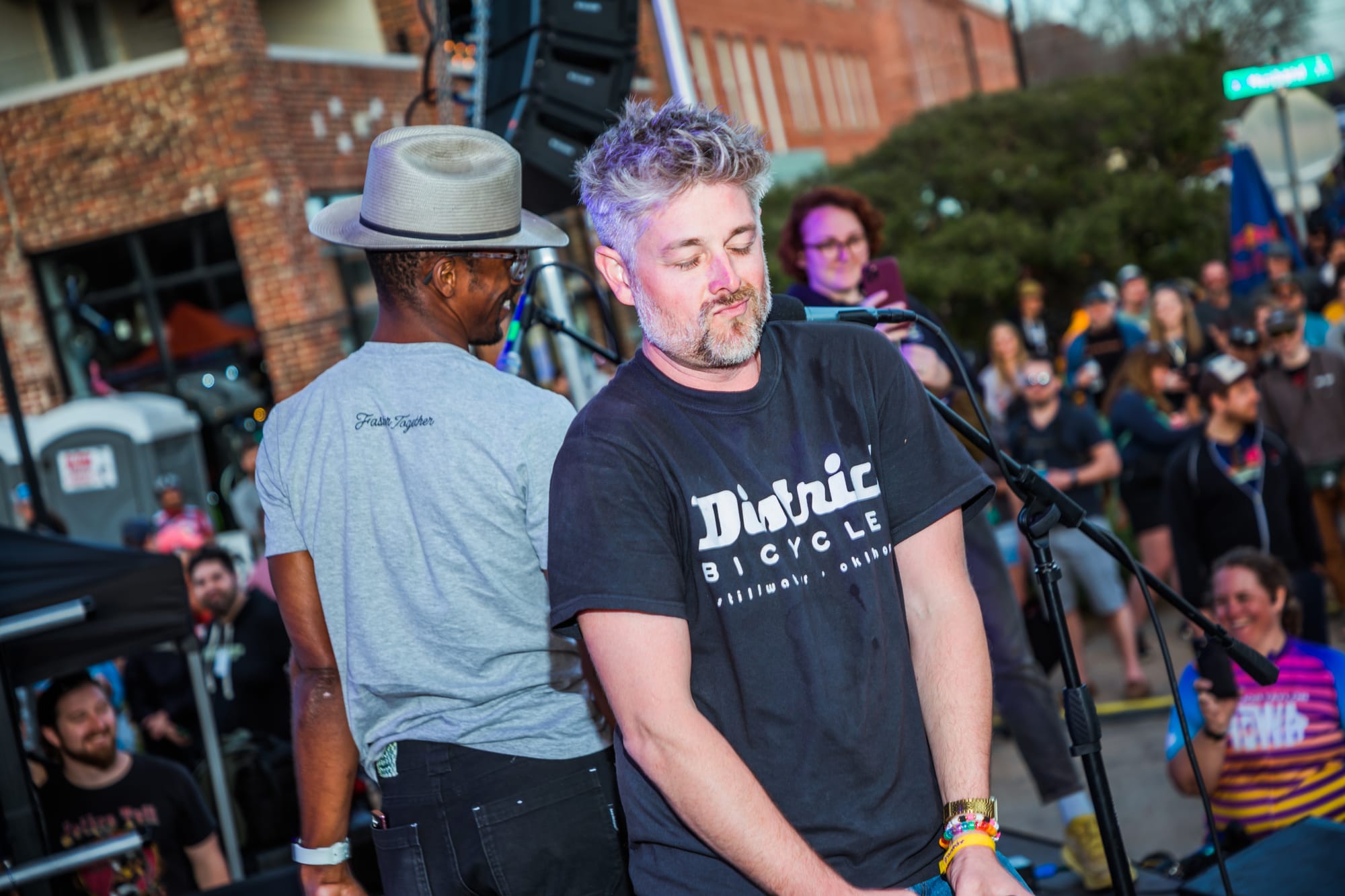 Bobby Wintle, wearing a District Bicycles T-shirt, dances at a microphone stand on an outdoor stage while a musician in a wide-brimmed hat stands behind him, with a crowd of festival attendees visible in the background.