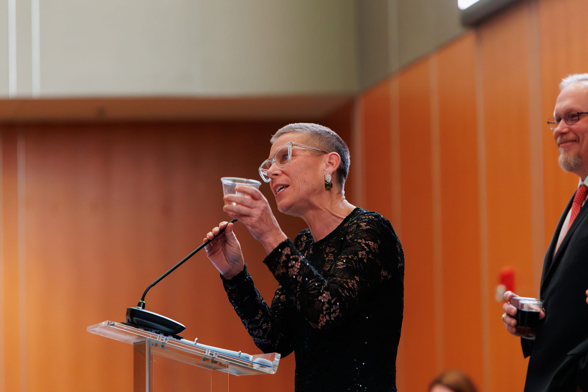 Alane Zannotti raises a toast at a clear podium during the Stillwater Chamber of Commerce Awards Gala. She wears a black lace dress and clear-framed glasses. 