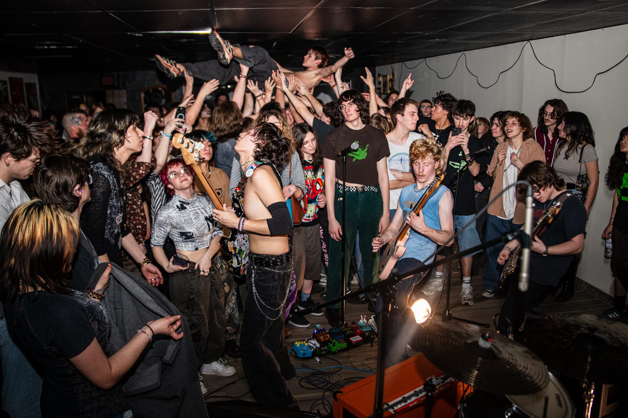 A crowd of young fans fills a small indoor venue, arms raised to support a shirtless crowd surfer overhead. Band members are visible performing at the right edge of the frame under stage lighting.