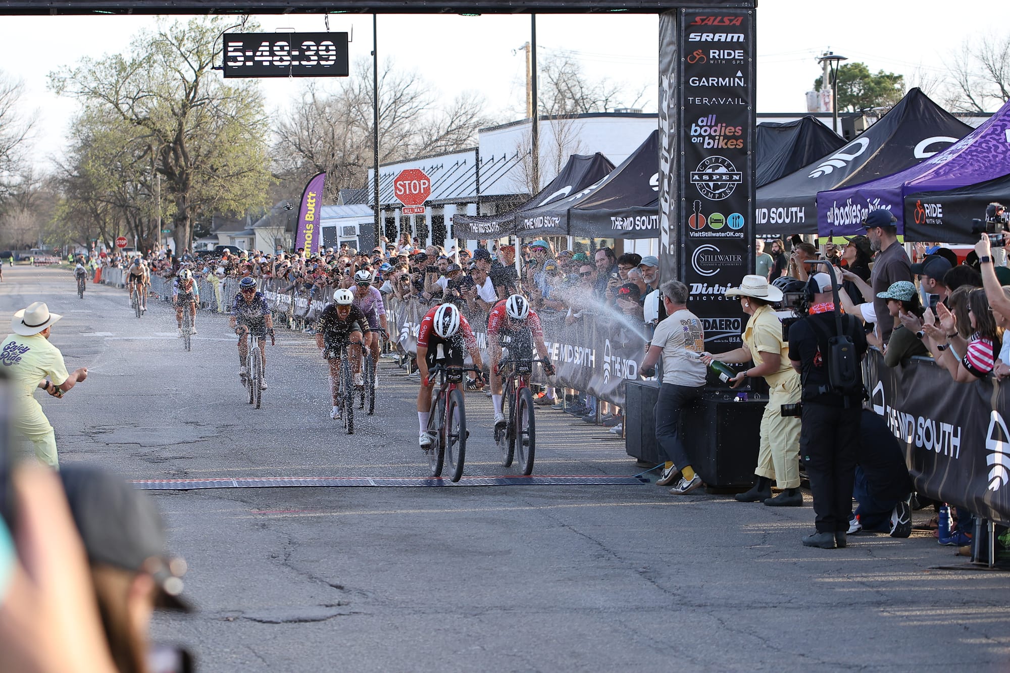 A nine-rider sprint group crosses the finish line at Block 34 in Stillwater as champagne sprays the chute during the women's pro race at the 2026 Mid South on Friday, March 13, 2026.